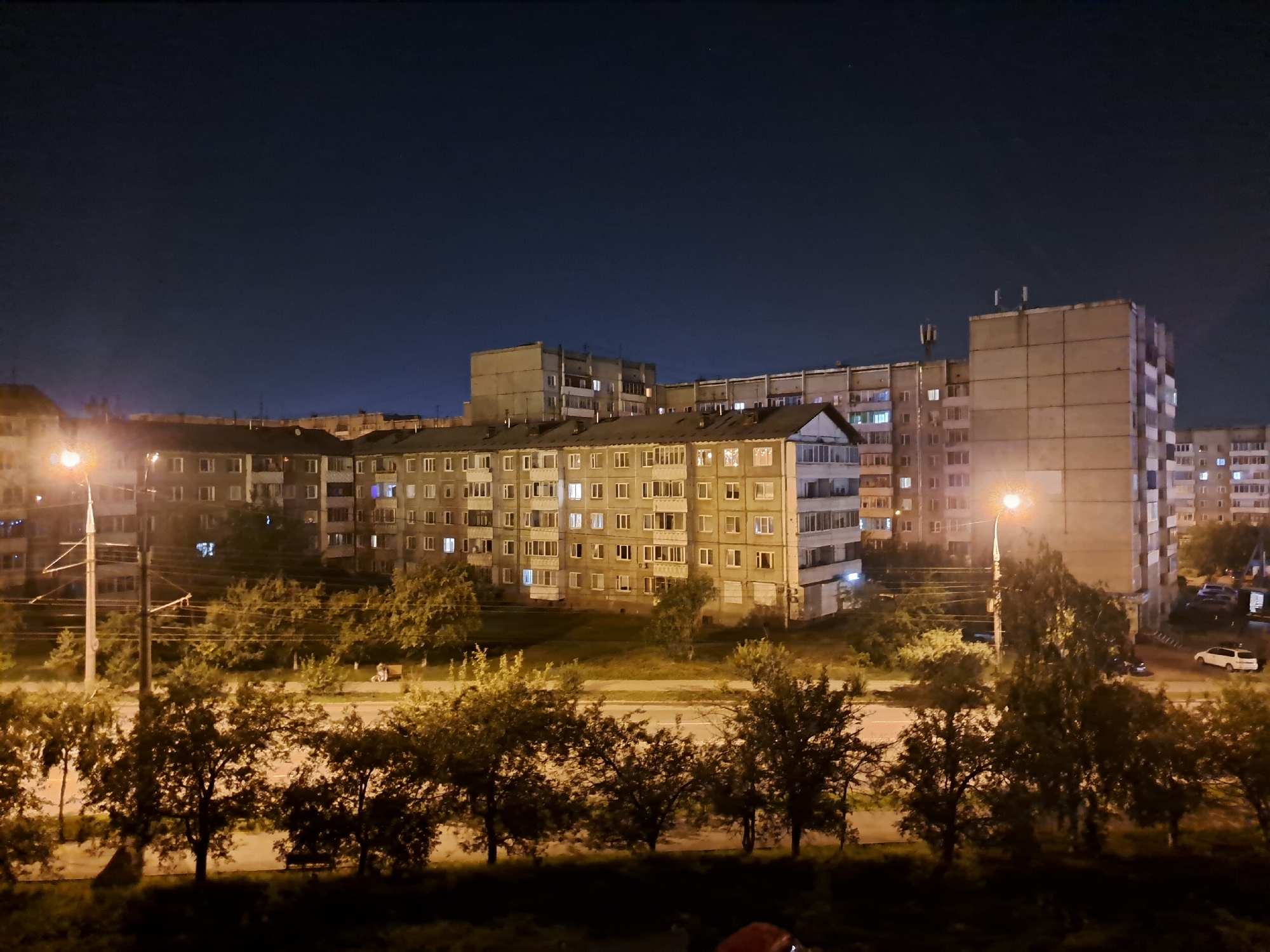 The image captures a nighttime view of a residential area in Irkutsk, Russia. The foreground is dominated by the dark silhouettes of trees, suggesting a park or a tree-lined street. Behind the trees, a road is visible with a streetlamp illuminating the area. Further back, several multi-story apartment buildings, characteristic of Soviet-era architecture, are lit from within, indicating occupancy. The sky is dark, with a few faint stars visible, and the overall atmosphere is that of a quiet urban night. A white car is parked on the right side of the image, and another car is visible further down the road. There are no discernible individuals in the frame. The scene appears to be a typical residential block at night, with artificial lighting from the buildings and streetlamps casting a warm glow on the surroundings.