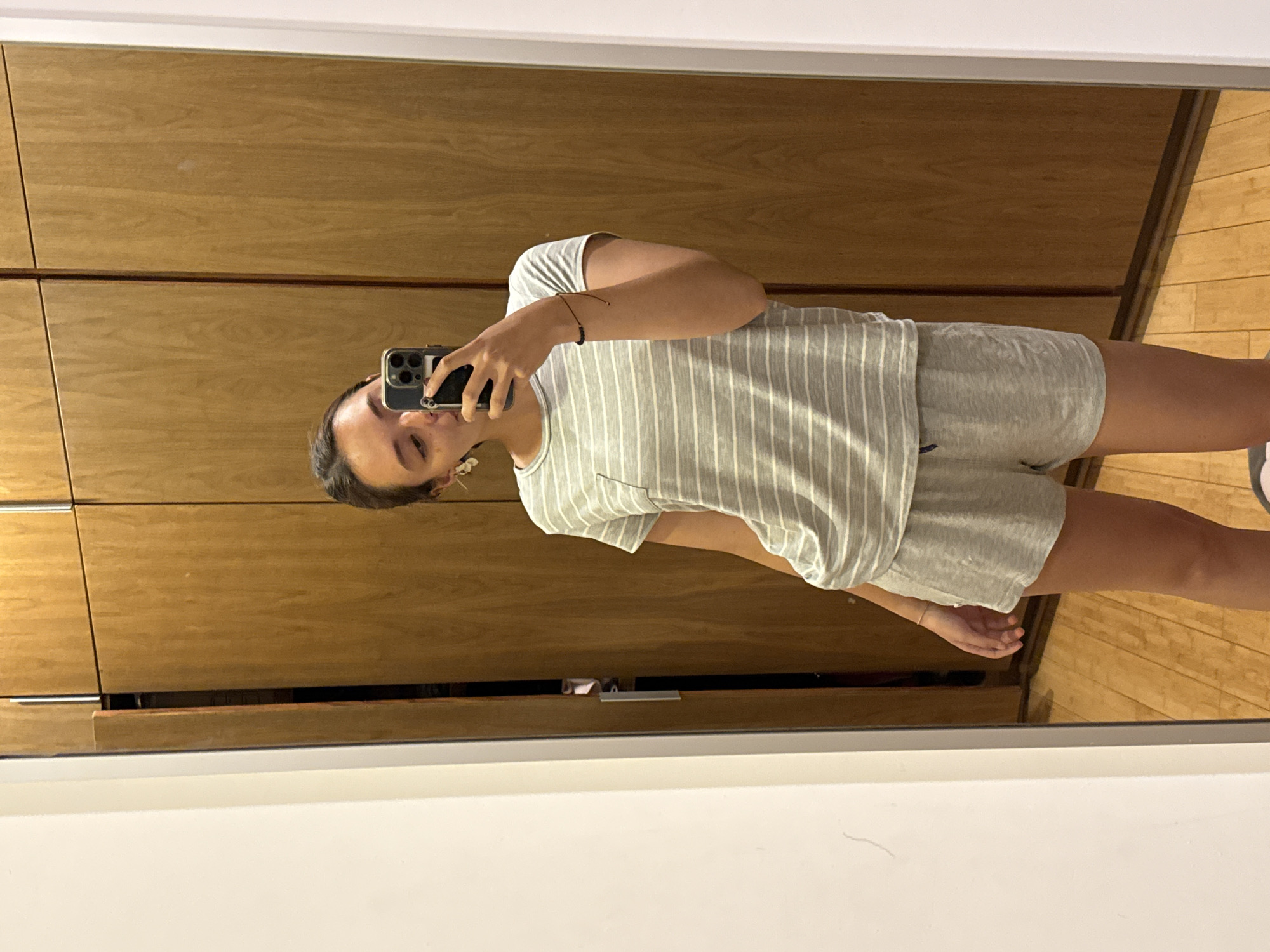 A person is taking a mirror selfie in front of a light wood paneled wardrobe. The person is wearing a gray and white striped t-shirt and gray shorts. The floor is made of light wood planks. The photo is framed by the white edge of the mirror. There is no evidence to suggest that the image was taken in San Bernardino Tlaxcalancingo, Mexico.