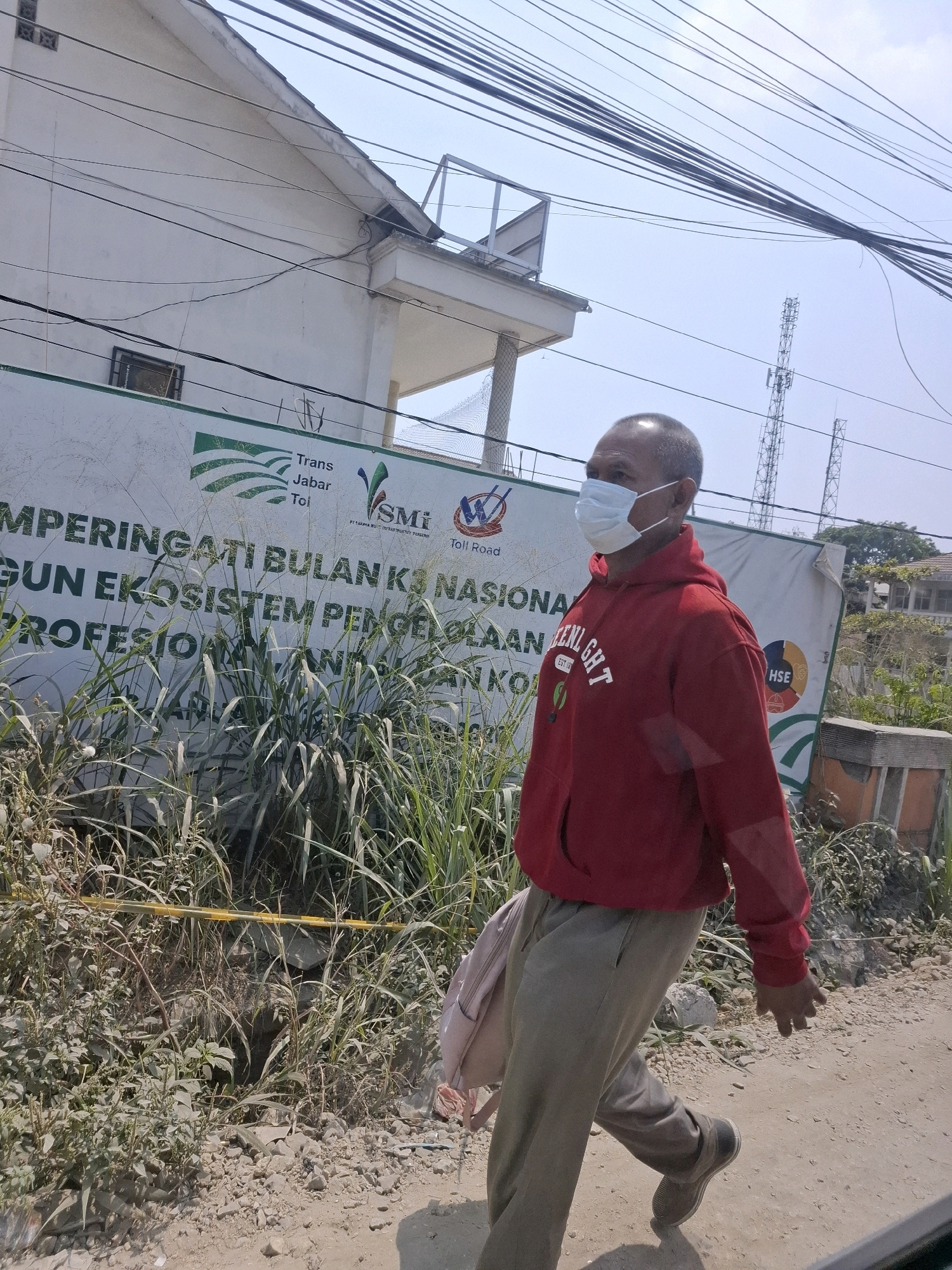 The image depicts an outdoor scene in Sukasari, Indonesia, under a bright, clear sky with a visible network of overhead wires. A man, wearing a light blue surgical mask, a red hooded sweatshirt with "GREEN LIGHT" printed on the chest, and grey pants, is walking to the right in the frame. He is carrying a light pink backpack over his left shoulder. He is walking on a dirt path next to a dense patch of tall green grass and weeds. Behind the vegetation is a white banner with green and blue text and logos, including "Trans", "Jabar", "Toi", "SMI", and "Toll Road". The banner appears to be an advertisement or announcement. In the background, there is a light-colored building and two cellular towers. The ground is rocky and dusty.