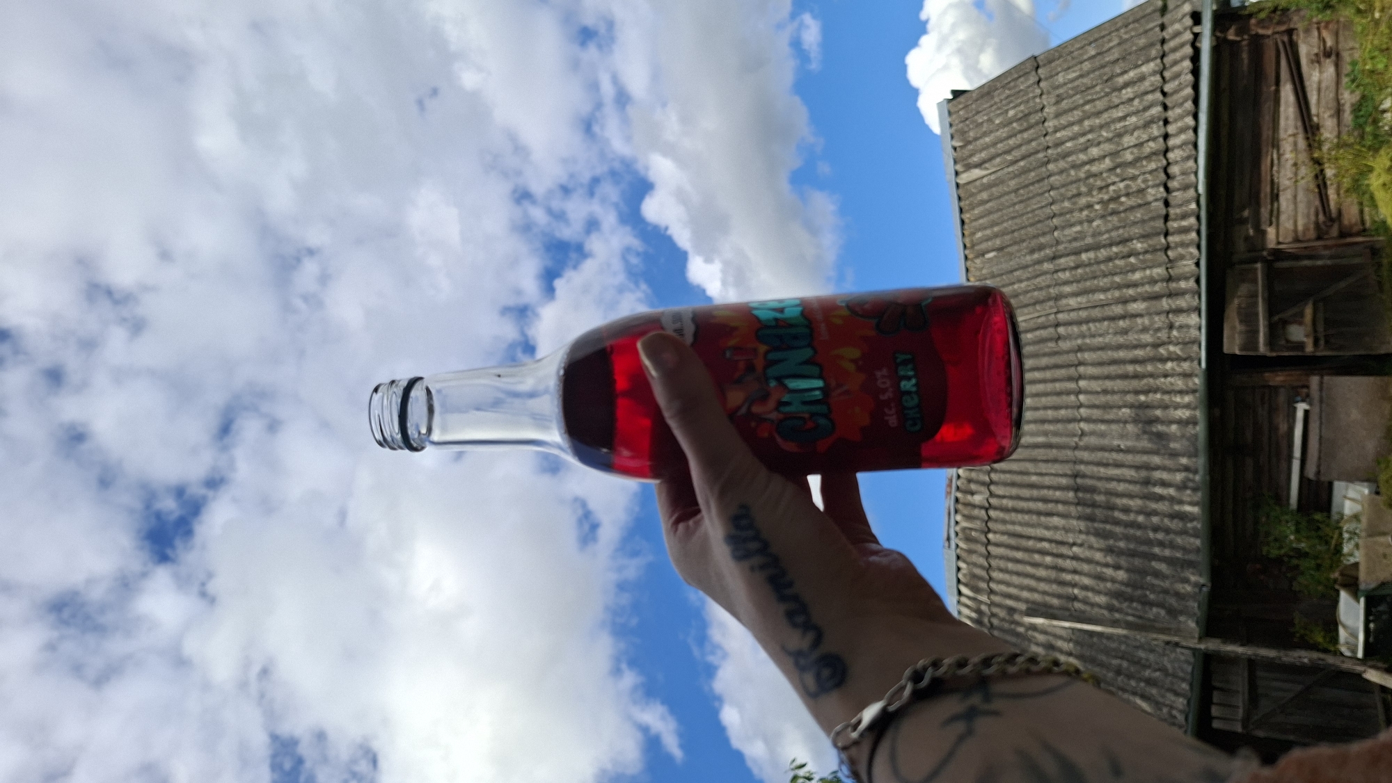 In Сваткаўскі сельскі Савет, Belarus, a hand with a tattoo reading "Kamilla" and a bracelet is holding a clear glass bottle filled with a red beverage. The bottle has a label that reads "CHINAZZI" and "CHERRY" with "alc. 5.0%" beneath it. The background shows a rustic wooden building with a corrugated roof against a blue sky with white clouds.