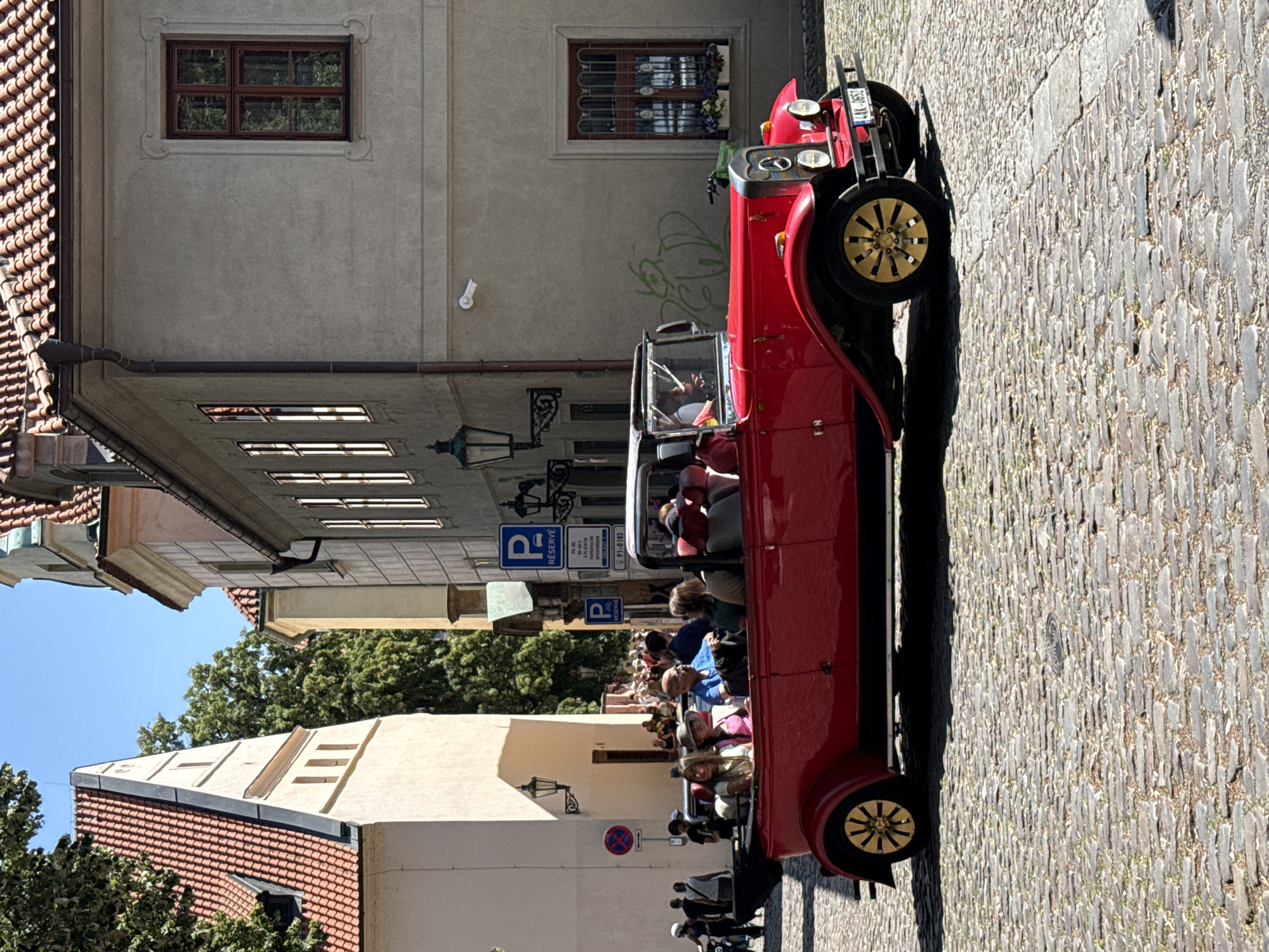 A red vintage car, likely a 1920s or 1930s convertible, is parked on a cobblestone street in Prague, Czechia. Several passengers, appearing to be tourists, are seated in the open-top vehicle, with some looking towards the camera. The car has yellow spoked wheels and a black interior. In the background, traditional European architecture with terracotta tiled roofs and muted-colored facades line the street. Two blue parking signs with the letter "P" are visible on poles. The sky is clear and blue, indicating a sunny day. The scene suggests a leisurely tour or a special event in a historic part of the city.