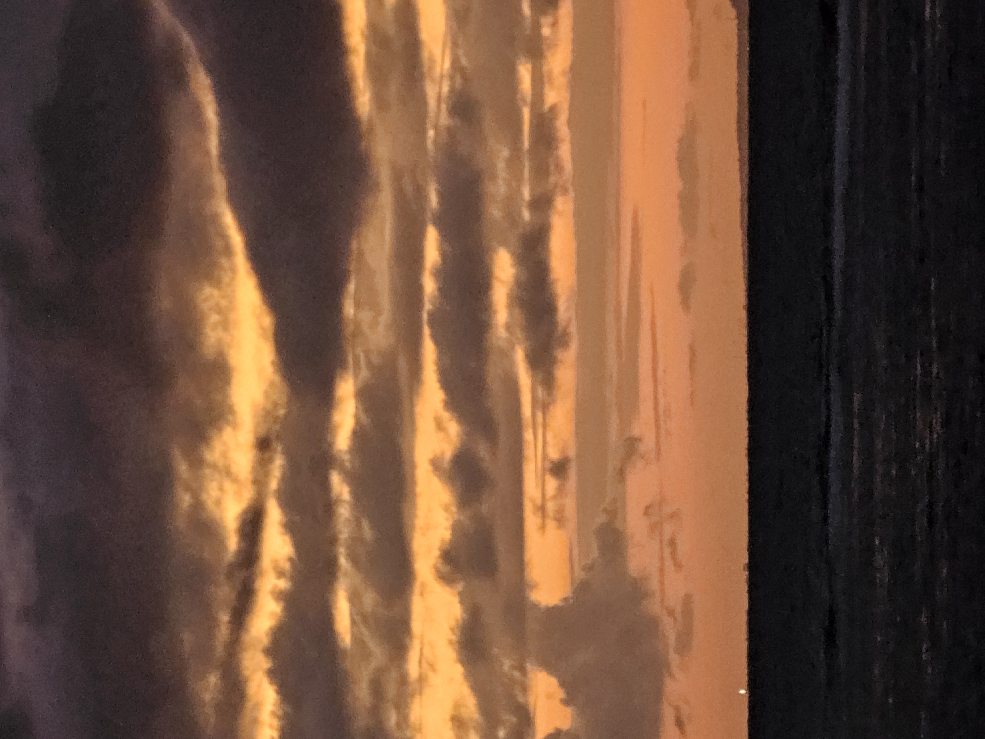 The image depicts a dusky sky over the ocean near Denpasar, Indonesia. The sky is filled with layered clouds, with hues of deep purple, orange, and yellow. The sun appears to have just set, casting a warm glow through the clouds. The ocean is dark and appears calm, with a thin, faint line of land visible on the horizon. There is a small, bright light on the horizon, possibly a distant ship or building. The overall scene is serene and atmospheric, evoking a sense of twilight over the sea.