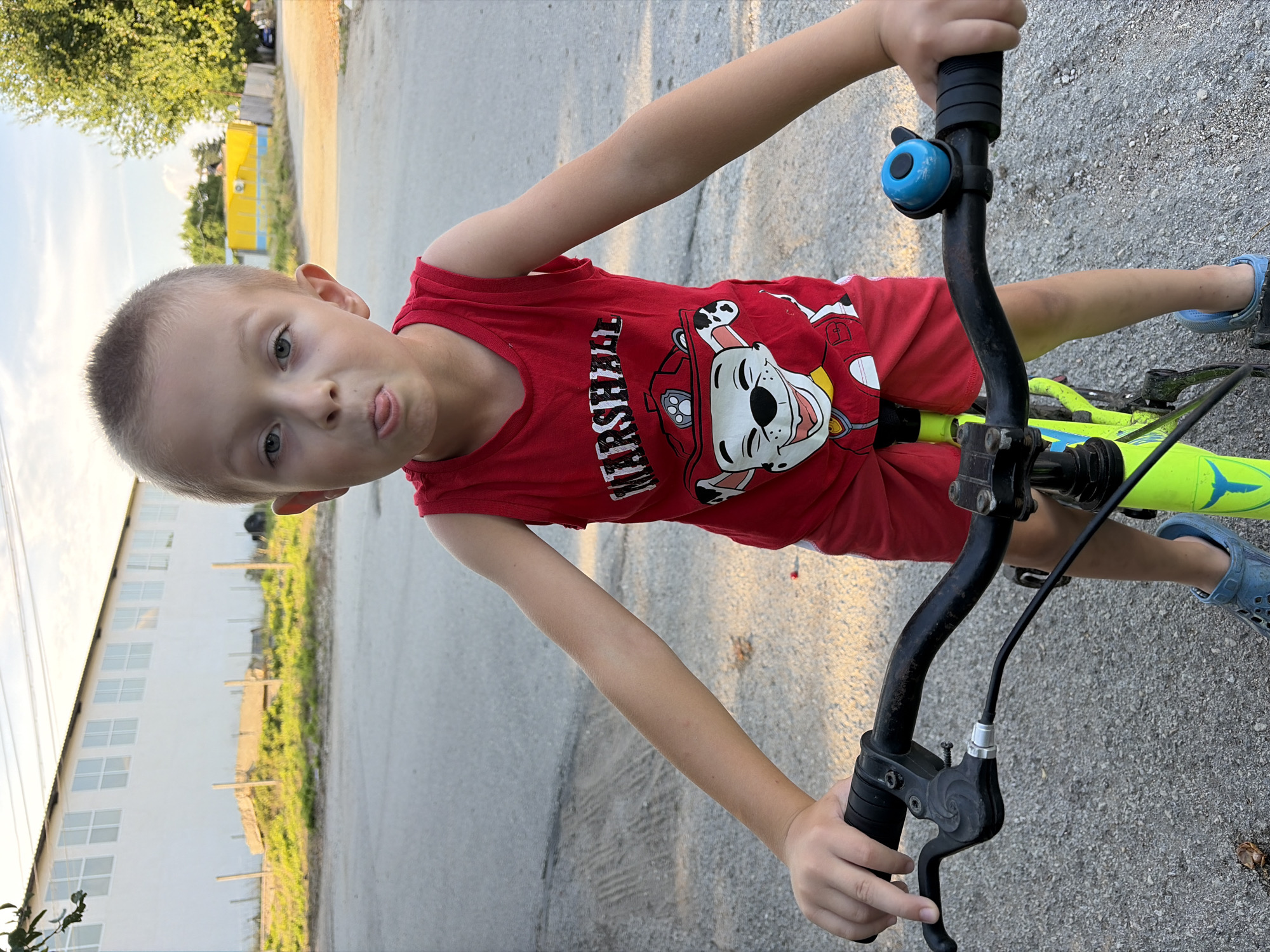 A young boy is standing on a street in Dolni Dabnik, Bulgaria, with his hands on the handlebars of a bright green bicycle. The boy is wearing a red tank top with a Paw Patrol character graphic and red shorts. He is sticking his tongue out at the camera. The street appears to be paved and is surrounded by a grassy area. In the background, there is a long, white building with many windows, a yellow building, and trees. The sky is partly cloudy.