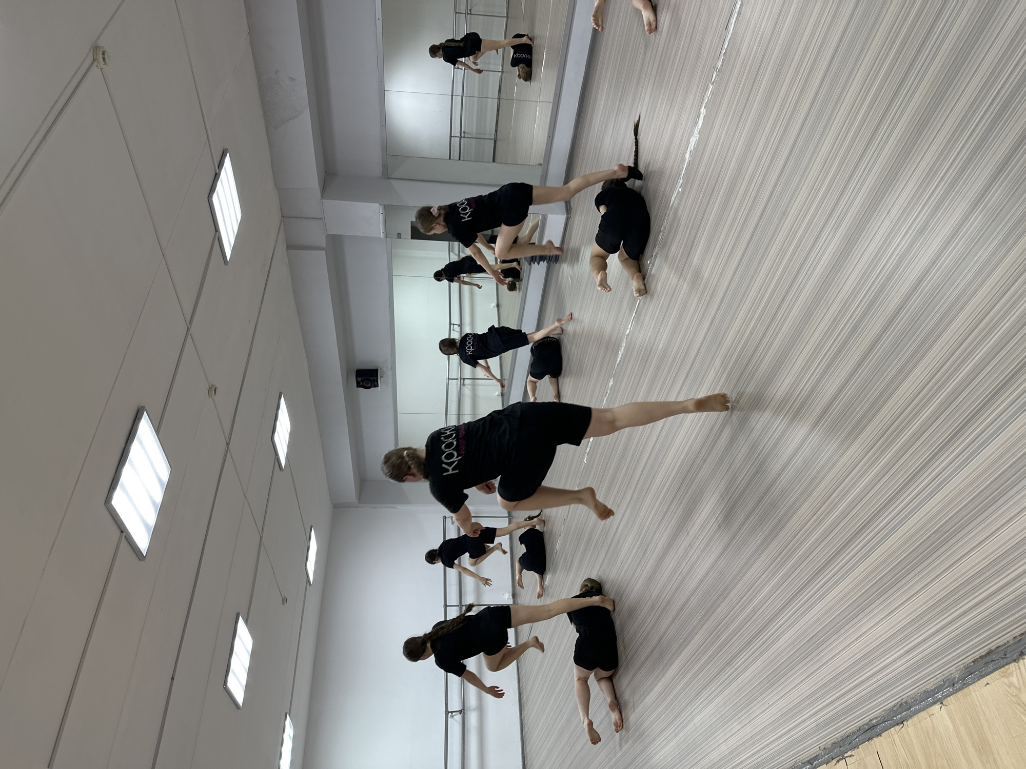 Inside a dance studio in Votkinsk, Russia, a group of dancers clad in black attire are practicing a routine. They are performing a coordinated move involving balancing on their feet while extending one leg horizontally. A large mirror on the wall reflects their actions, creating a sense of depth and repetition. The studio features a light-colored wooden floor and a wall with square panel lights. The scene is well-lit and captures a moment of focus and physical exertion.