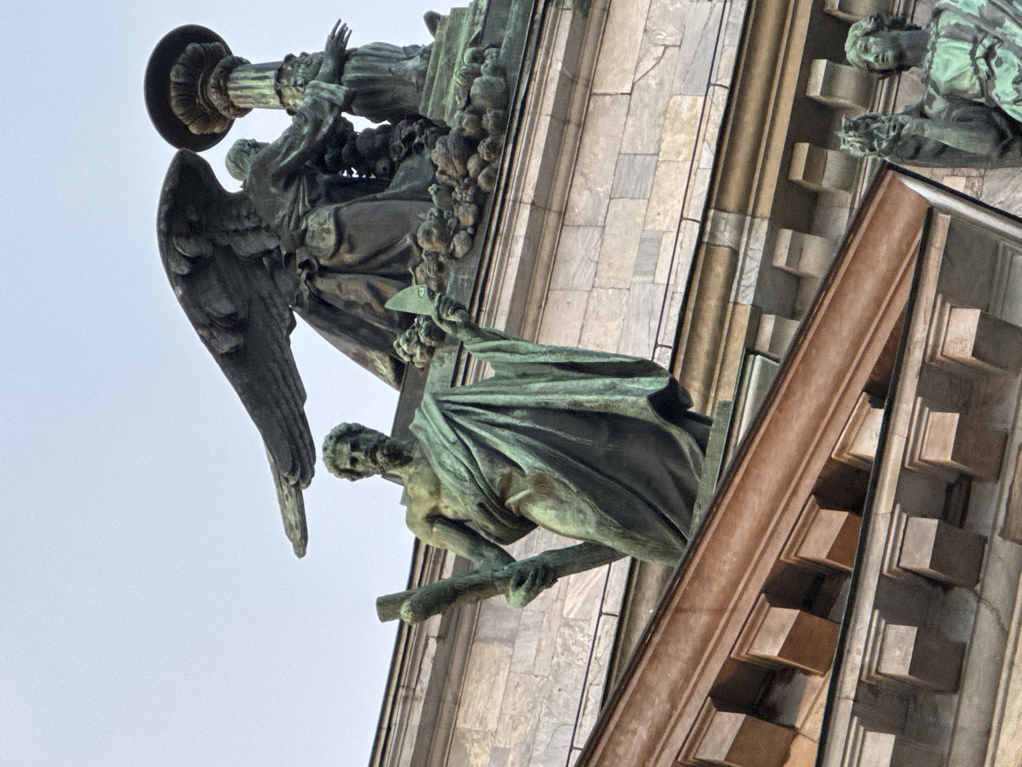 This image shows architectural details and sculptures atop a grand building. In the foreground, a bronze statue of a man with a beard, draped in a toga, holds a staff. Behind him and to the right, a large bronze angel with outstretched wings appears to be tending to a candelabra. Further to the right, another figure, partially visible, is also part of the sculptural decoration. The building itself is constructed with stone blocks and features ornate cornices and decorative elements. The sky is overcast. This is a view of the sculptures and architectural features adorning the dome of Saint Isaac's Cathedral in Saint Petersburg, Russia.