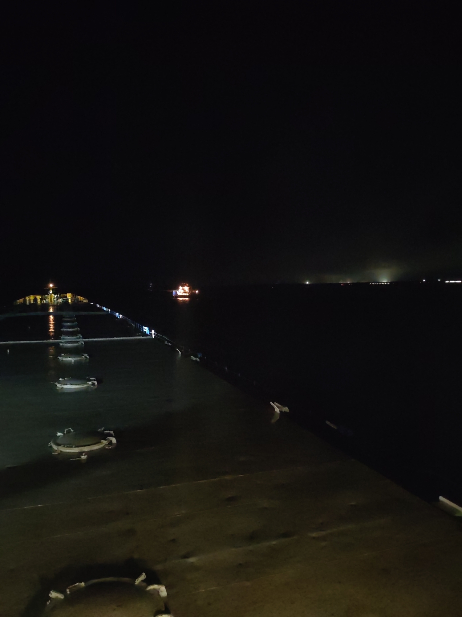 The image shows a night view from a ship at sea. In the distance, there are lights from what appear to be other ships and a coastline. The foreground depicts the deck of the ship with several circular objects, possibly hatches. The lighting is dim, with artificial lights creating reflections on the wet surfaces of the deck.