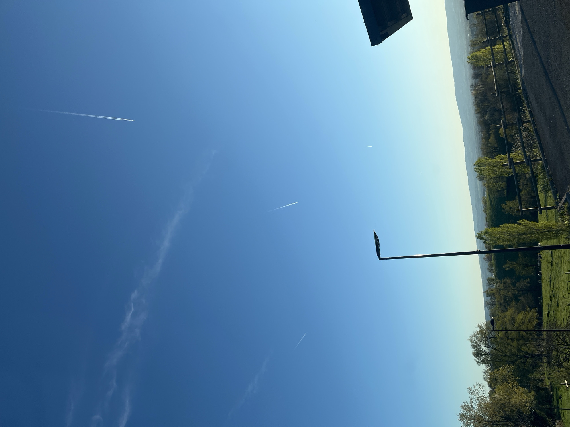 The image shows a daytime scene in Chambœuf, France. The sky is predominantly blue with contrails from airplanes visible. On the right side of the frame, a portion of the landscape is visible, including trees, a field, and a fence. A street light pole is also present. The perspective of the image is rotated, as if viewed on its side.