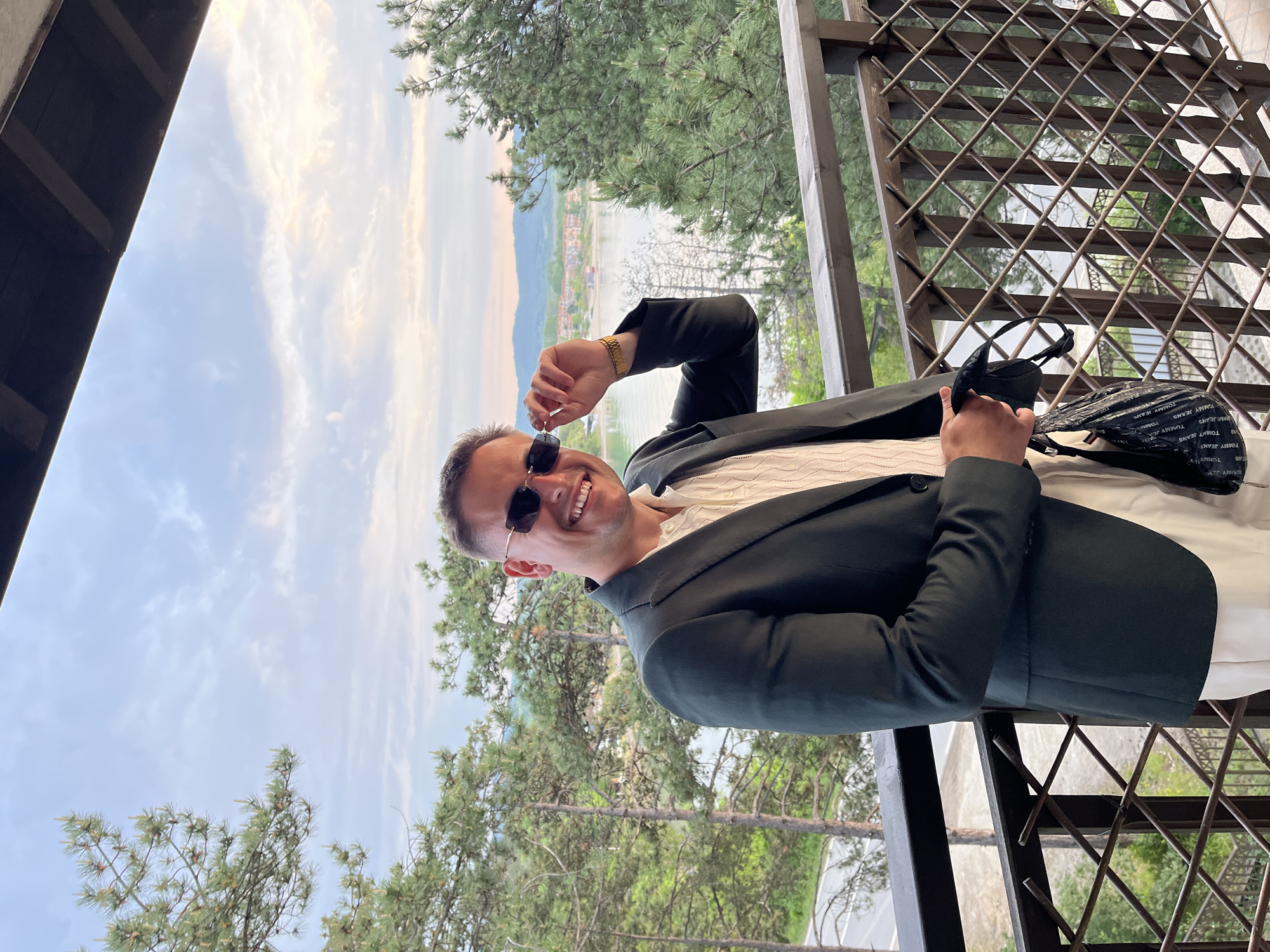 A man is smiling and wearing sunglasses and a dark suit. The location appears to be an elevated outdoor area with a lattice railing. In the background, there is a view of a body of water, trees, and possibly a settlement. This is likely Sozopol, Bulgaria, given the request. The man is holding his sunglasses in one hand and a black bag with white lettering in the other.