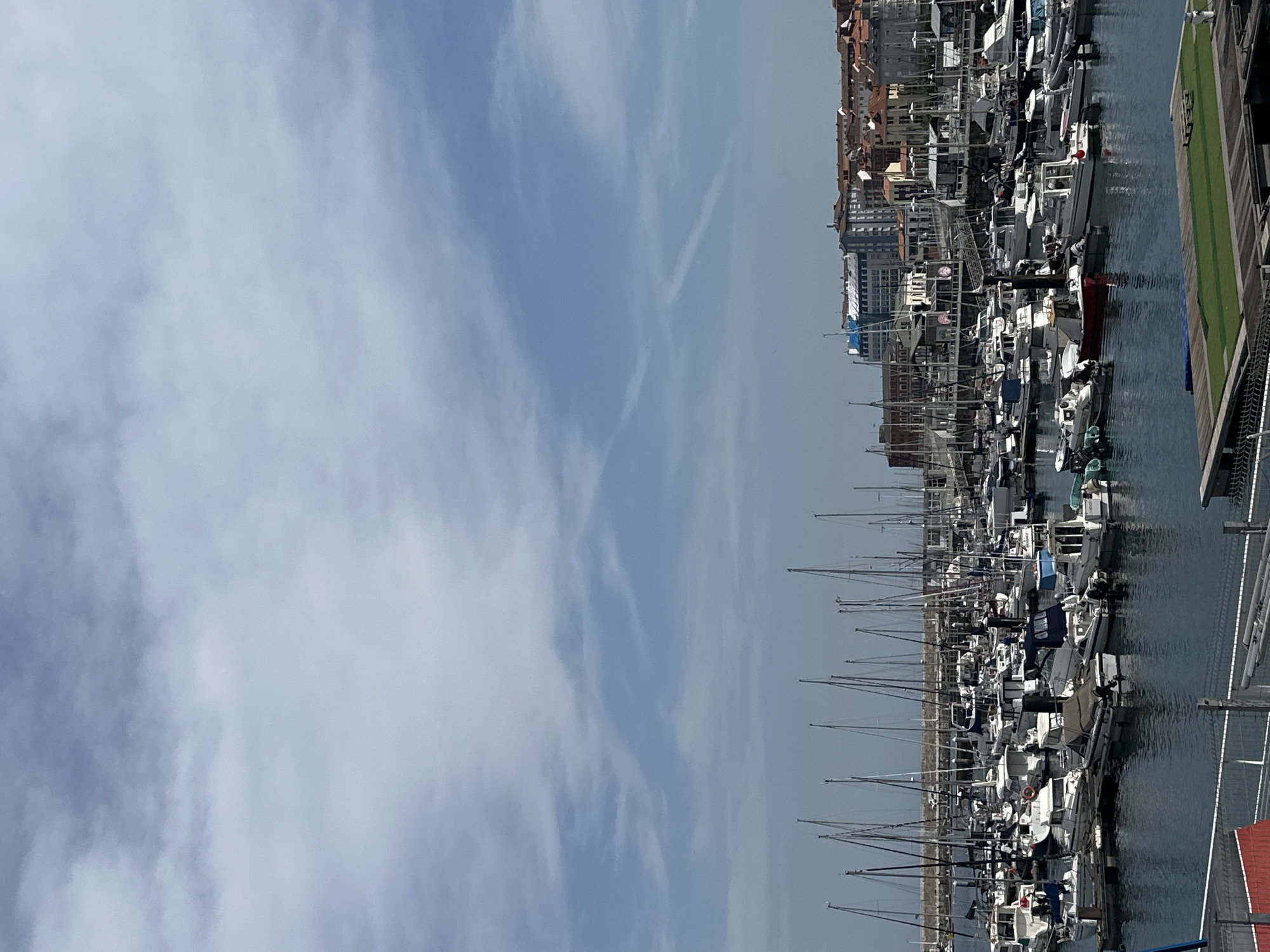 The image depicts a marina in Gijón/Xixón, Spain, with numerous sailboats and yachts docked along piers. The vessels are tightly packed, displaying an array of white hulls and masts. In the background, buildings rise from the shore, showcasing a mix of traditional red-tiled roofs and modern architecture. The sky is partly cloudy, with varying shades of blue. A section of pier, possibly wooden, is visible on the right side of the image. The water is calm, reflecting the boats and surrounding structures.