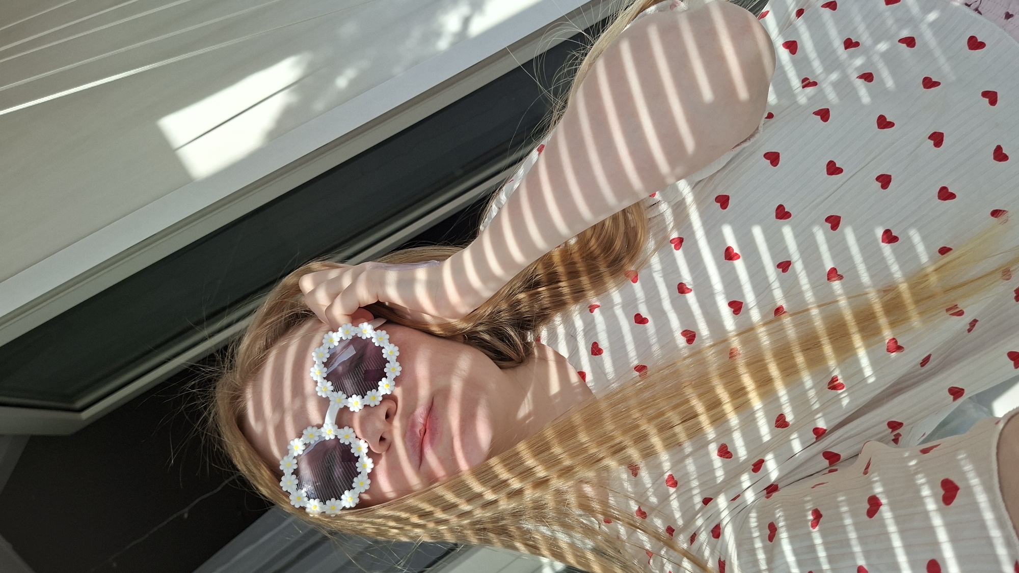 A blonde girl wearing flower-shaped sunglasses lies on a white surface patterned with red hearts. Sunlight casts striped shadows across her face and body. She is holding the sunglasses with her hand. The location is most likely indoors, near a window. The location in the city of Chaikovsky, Russia, cannot be verified from the image alone.