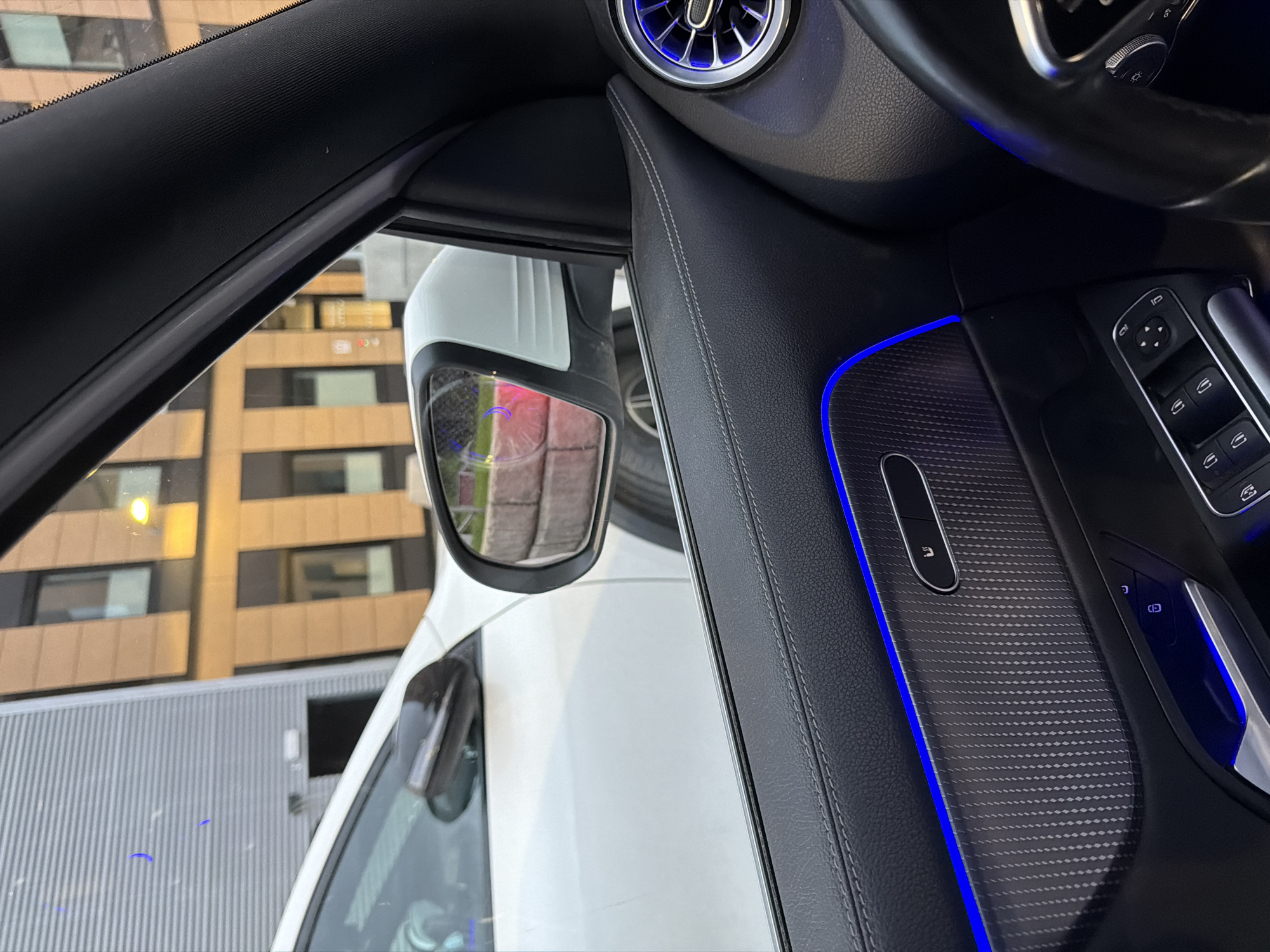 The image provides an interior view from a car parked in Harstad, Norway, likely in a residential or commercial area judging by the modern building exterior visible through the car window. The building features a textured grey facade on the left and a beige facade with dark rectangular window frames on the right, illuminated by warm yellow lights.
The car's interior is visible in the foreground, showcasing a black leather door panel with contrasting white stitching. A carbon fiber-like trim piece is integrated into the panel, accentuated by a vibrant blue ambient light strip above it. Below this, a chrome door handle is partially visible, along with a row of window control buttons. A circular air vent with a blue illuminated ring is situated higher on the door panel. The steering wheel, also black, is partially visible on the right side of the frame, with some control buttons.
The car's side mirror reflects a glimpse of the exterior environment, showing a portion of a white vehicle parked next to it, and a wall constructed of beige bricks. The overall scene suggests an evening or twilight setting, given the artificial lighting. There are no people visible in the image.