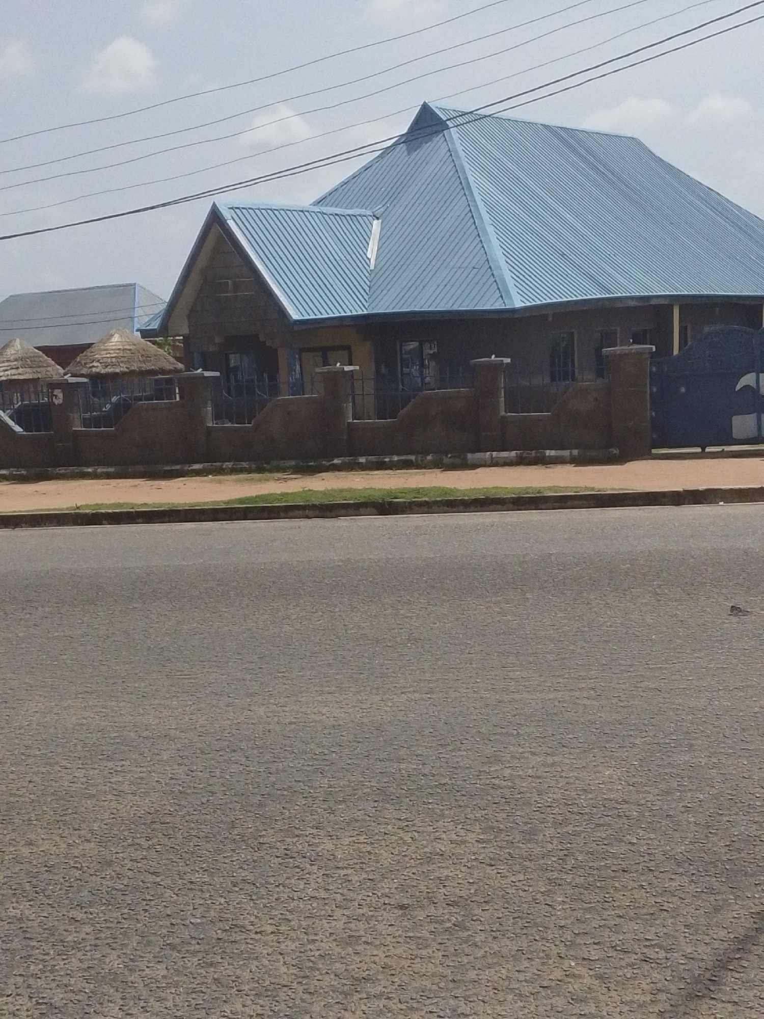 This is an outdoor, eye-level, medium shot of a building with a blue, corrugated metal roof in Jalingo, Nigeria. The building has a stone facade and several windows. In front of the building is a low wall with pillars, and a dark metal gate is visible on the right side of the frame. In the foreground, a paved road runs horizontally across the image. The sky is overcast with some clouds.