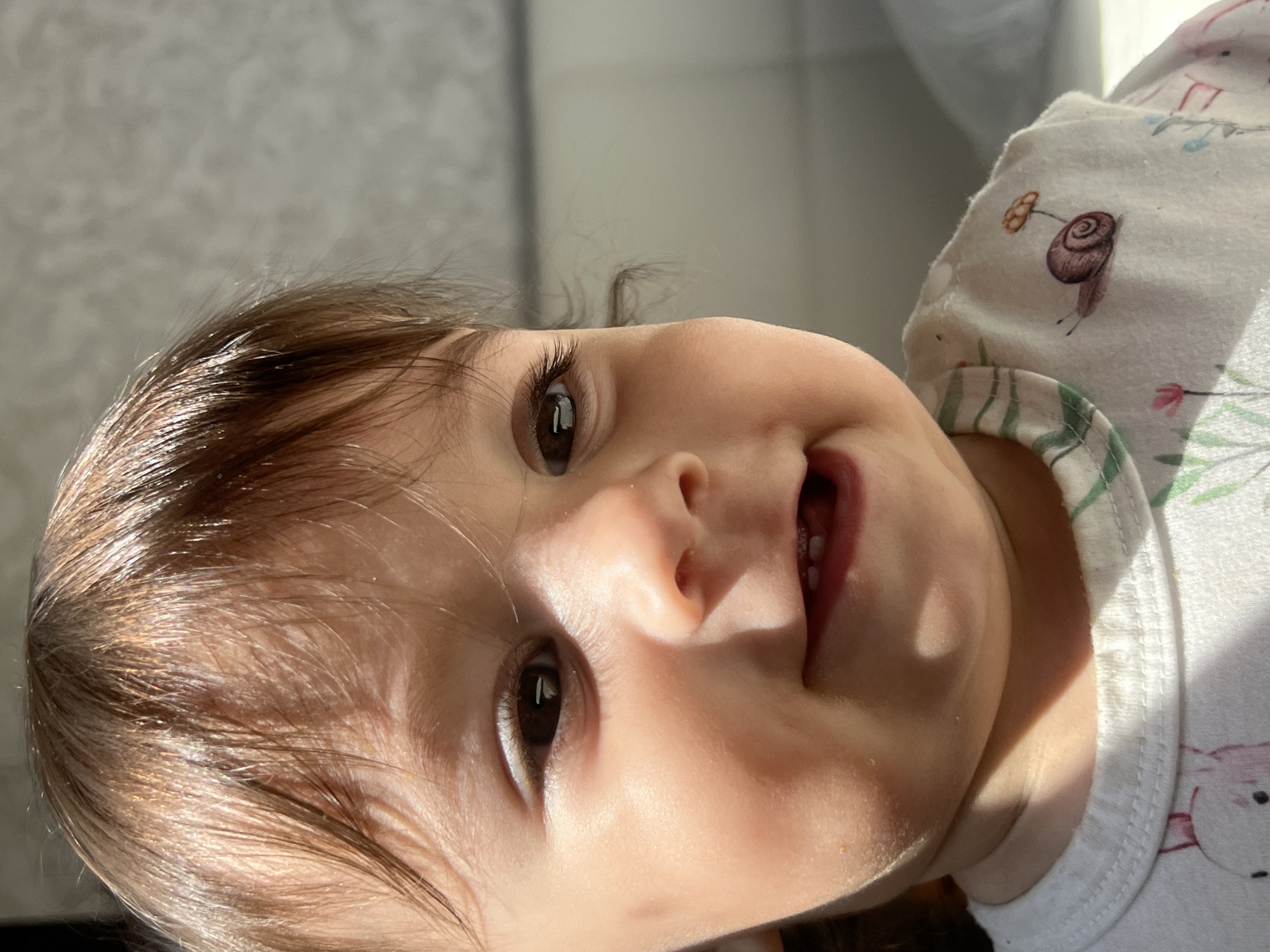 A close-up portrait shows a young child or baby, likely an infant or toddler, looking directly at the camera with a gentle, curious expression and a slight smile. The child's head is tilted sideways to the left, as the image is rotated 90 degrees clockwise. The child has dark brown hair, which appears soft and fine, with prominent highlights from natural sunlight. Large, dark brown eyes with long eyelashes are open wide. Two small lower teeth are visible as the child smiles.
The child is wearing a light-colored, possibly white, garment, which appears to be a top or onesie. The fabric is patterned with small, delicate illustrations, including a distinct brown snail with an orange mushroom-like growth next to it, as well as faint floral or plant motifs in green, pink, and blue. A green and white striped trim is visible around the collar area.
Strong natural light, likely sunlight, illuminates the child's face, hair, and clothing, creating bright highlights on the skin and hair, and casting soft shadows that add depth to the features. The background is softly blurred, appearing as a light grey or off-white surface, suggesting an indoor setting bathed in daylight. The overall mood is warm, engaging, and joyful. The photo was taken in Elena, Bulgaria.