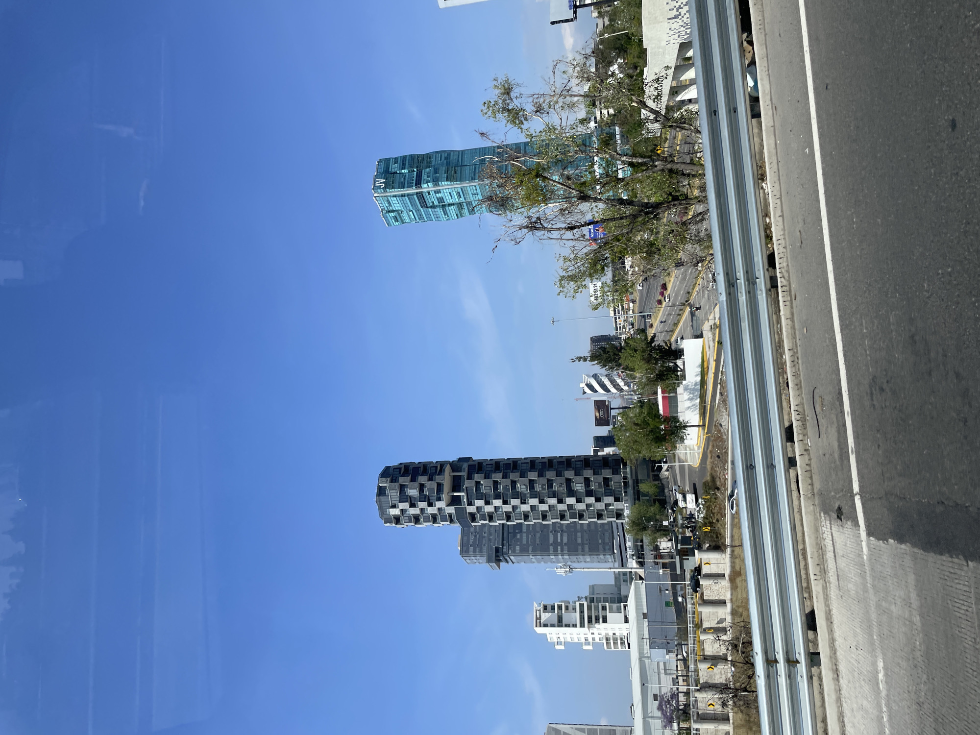 The image shows a view of a modern urban landscape in San Bernardino Tlaxcalancingo, Mexico. The perspective is from a high vantage point, likely a bridge or elevated highway. A guardrail runs along the right side of the frame, with the road surface visible beyond. Several high-rise buildings dominate the scene, set against a clear blue sky. The first building is a multi-story structure with a black and grey patterned facade. Further in the background, there is another modern-style skyscraper with light blue glass, partially obscured by bare tree branches. Smaller buildings, roads, and some greenery are visible between the high-rises.
