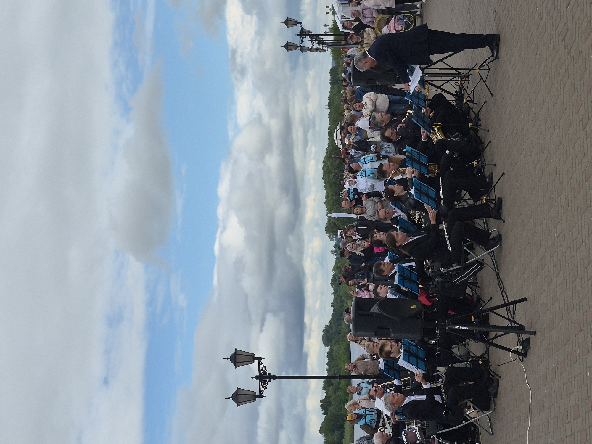 An outdoor musical performance by a brass and woodwind band is depicted on a paved promenade adjacent to a wide river in Sarapul, Russia. The event takes place under a sky largely covered by stratocumulus clouds, with some clear blue patches. In the foreground, approximately 15 to 20 musicians, predominantly adult males, are seated on folding chairs, performing with various instruments including saxophones, clarinets, flutes, trombones, trumpets, and low brass instruments. Each musician has a blue music stand with sheet music. An adult male conductor, dressed in a dark suit, stands in front of the band, facing them, and directs the performance. Two large black PA speakers on tripods are positioned among the musicians, with power cables extending across the reddish-brown paver stone surface. Ornate black streetlights with dual lanterns are prominent on the left side of the image, and a series of similar lights extends into the background on the right. Behind the band, a casual crowd of spectators of various ages observes the performance. The background features a wide body of water, identifiable as the Kama River, with a densely forested bank on the opposite side. The setting is consistent with the Sarapul Embankment (Набережная Сарапула) in Sarapul, Udmurt Republic.