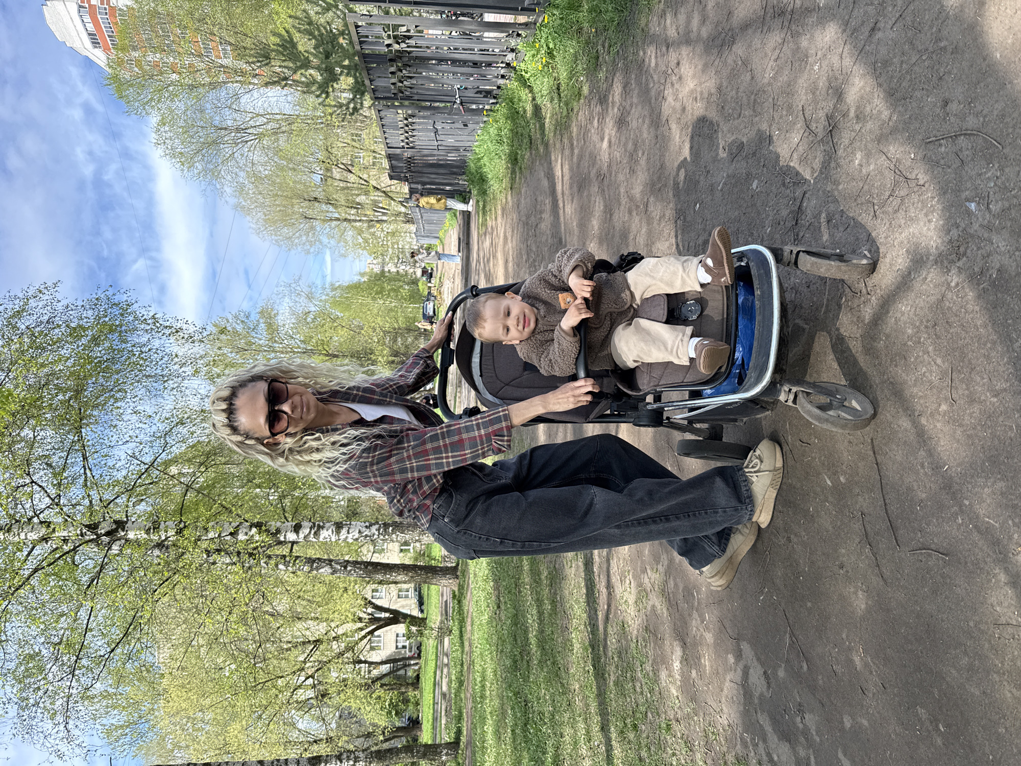 A woman with long blonde curly hair, wearing sunglasses, a plaid shirt, dark jeans, and sneakers, is standing next to a blue baby stroller on a dirt path. A baby, wearing a brown sweater and beige pants, is sitting in the stroller. The woman has her hands on the stroller. The scene is outdoors, with trees and grass visible in the background. A black metal fence runs along the edge of the path. A multi-story building can be seen in the distance. The photo was taken at Rusanovo, Russia.