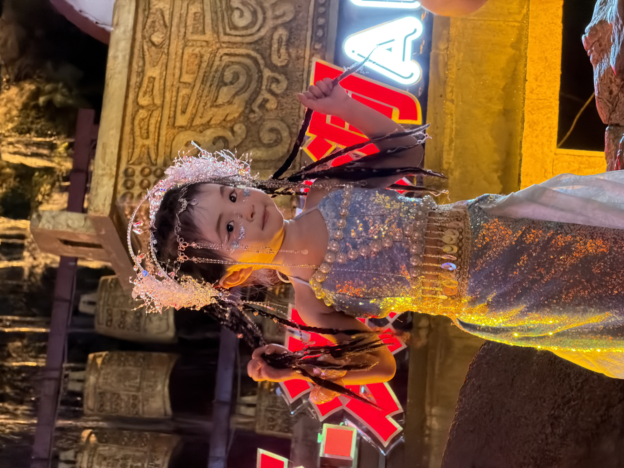 A young child, adorned in a sparkling blue and silver dress with pearl embellishments and a jeweled headdress, stands on a dimly lit stage in Yuzhong District, China. The child is holding their braided hair, which is decorated with strands of beads and metallic accents. The background features large golden bells on a wooden rack and a neon sign with the letter "A" illuminated in white. The stage setting suggests a cultural performance or festival.