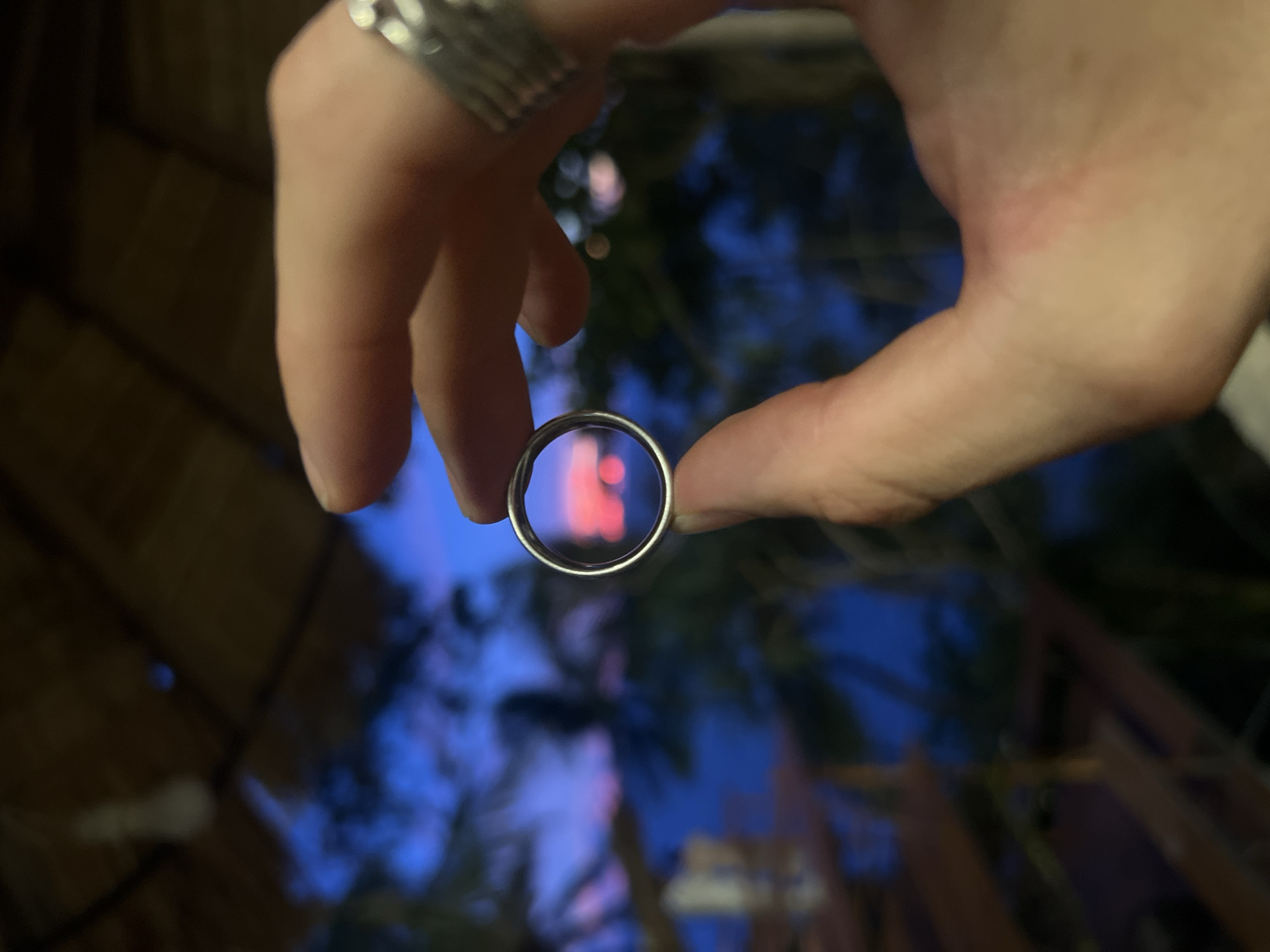 A person's hand, adorned with a silver ring on their index finger, holds a silver ring in front of their palm, framing a blurry background. Through the inner circumference of the held ring, a distorted reflection is visible. It appears to be the outlines of bright red or pink lights against a deep blueish-purple background, possibly indicative of the sky at dusk or illuminated elements within a scene. The background is generally out of focus, with hints of dark foliage and wooden structures, suggesting an outdoor setting, potentially a tropical environment given the location context of Karon, Thailand. The lighting suggests it is either twilight or nighttime, with artificial lights contributing to the color of the background. The overall impression is one of a casual moment, perhaps capturing a decorative element or a specific view through the ring.