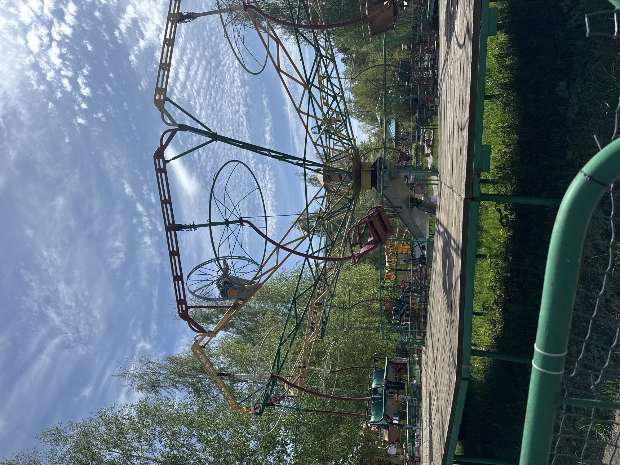 The image depicts a children's carousel in the city of Chaikovsky, Russia. The carousel is made of metal and painted in various colors, including red, yellow, and green. It has seats hanging from it, some of which are visible. The carousel is located on a wooden platform surrounded by green grass and trees. The sky above is blue with some white clouds. A green fence is in the foreground on the right.