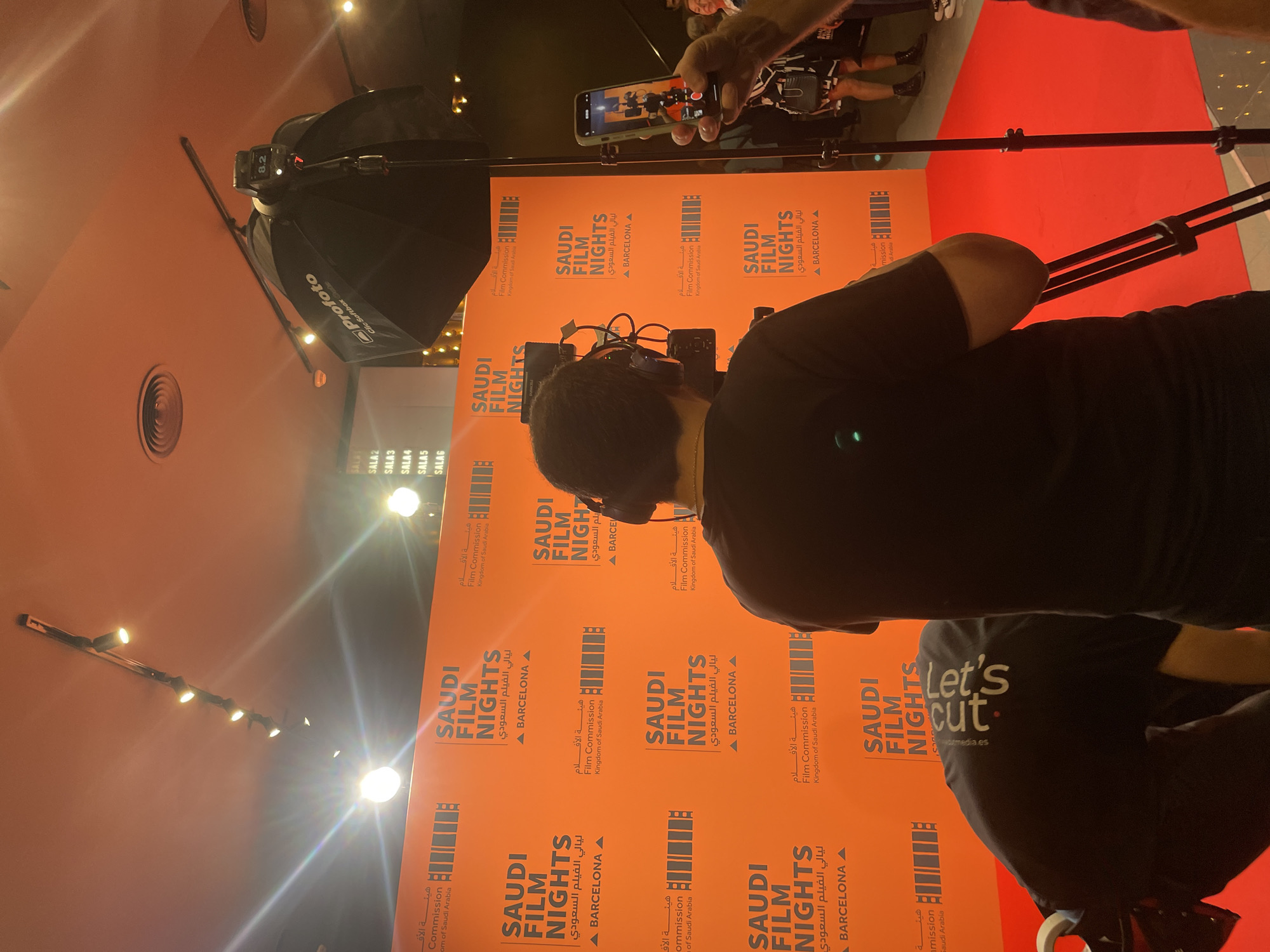 The image shows a behind-the-scenes moment at what appears to be a film event or promotion called "Saudi Film Nights" in Barcelona, Spain. The event features an orange backdrop with the event's name and logo in both Arabic and English. A photographer is capturing the scene with a professional camera setup, including a large "Profoto" brand softbox lighting. An individual is recording the event using a smartphone. A person wearing a black t-shirt with "Let's cut" and the company's media es. can be seen from behind. The event takes place in a venue with a red carpet. Several lights can be seen on the ceiling.