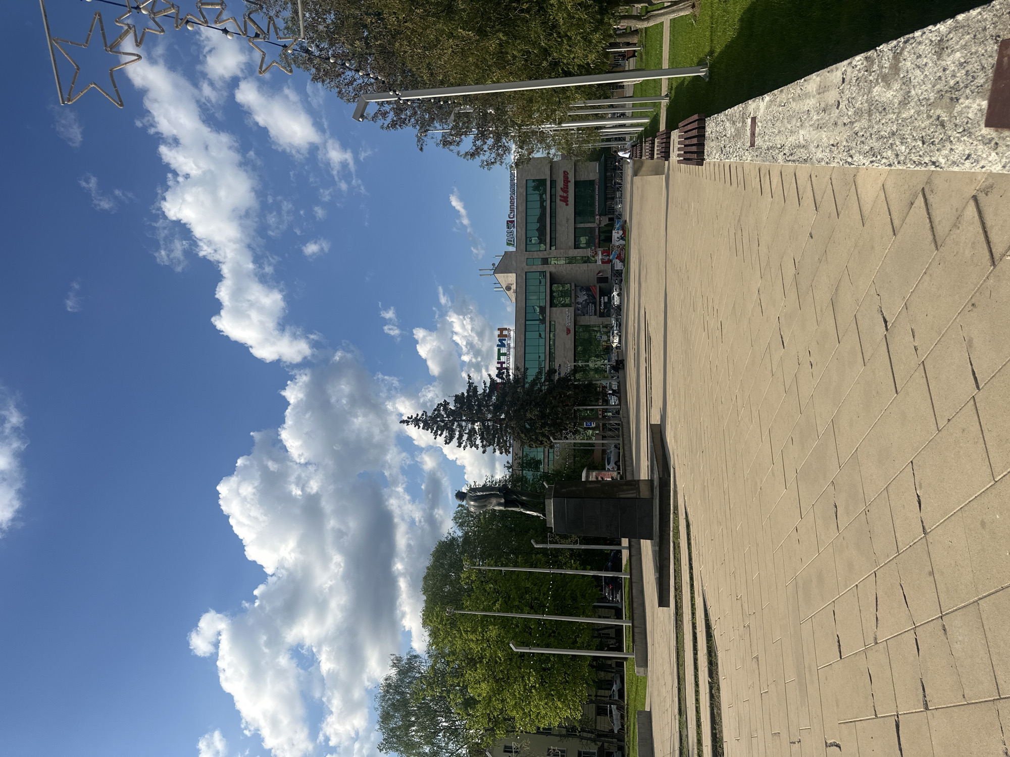 The image shows a view of Naro-Fominsk, Russia, on a sunny day. A paved pedestrian area leads toward a building with multiple logos and signs in Cyrillic, including "Мясное супермаркет." Trees are visible along the walkway, as well as a statue on a pedestal. The sky is blue with cumulus clouds.