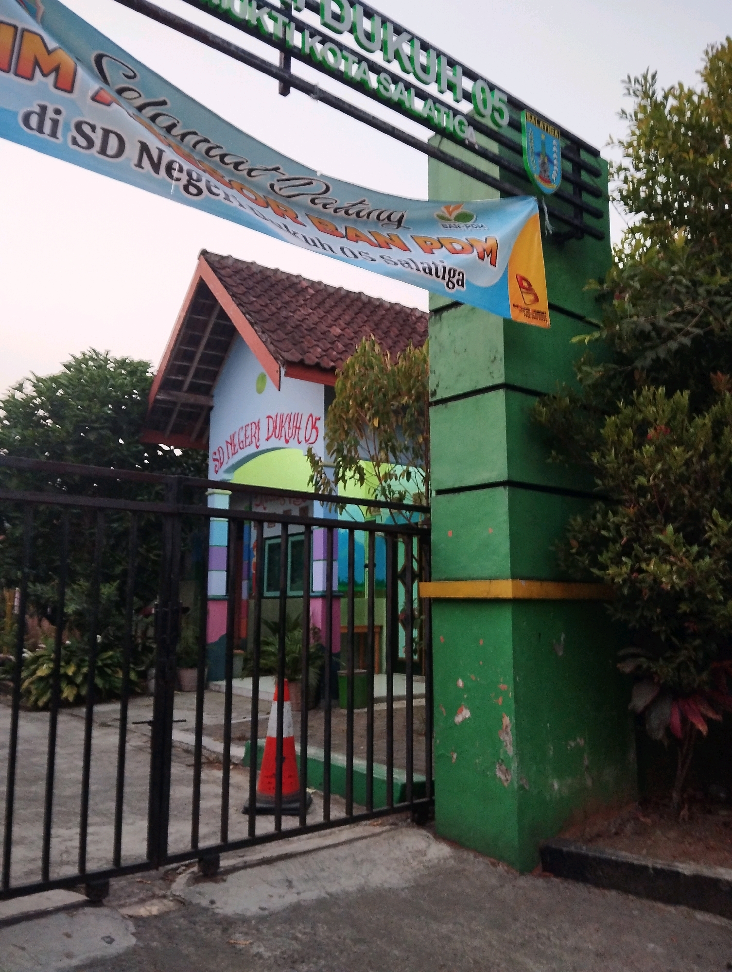The image captures the entrance gate and a portion of a building at SD Negeri Dukuh 05, located in Salatiga, Indonesia. A large blue and white banner hangs across the entrance, with text that appears to read "SELAMAT DATANG di SD NEGERI DUKUH 05 SALATIGA". The building, painted with various bright colors, has a distinctive tiled roof. In front of the building, behind a black metal fence, is an orange and white traffic cone. The entrance pillar is painted green with a yellow band. Lush green trees and foliage are visible on the right side of the image. The sky is overcast. The overall scene depicts a public institution, likely a primary school, with a welcoming banner.