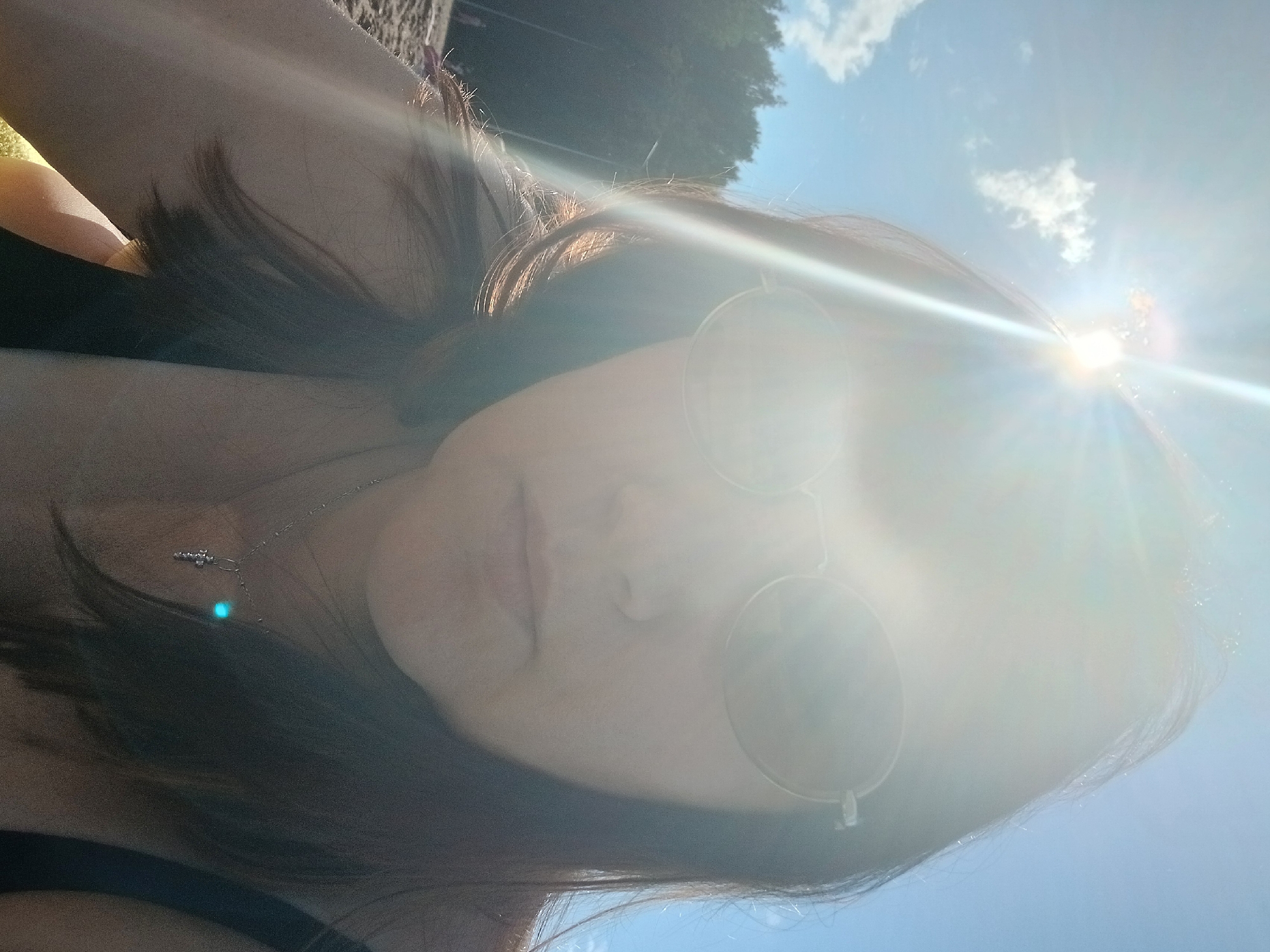 The media file is a selfie taken outdoors on a sunny day. The subject is a person, likely a woman, with dark hair, wearing sunglasses. The sun is bright, creating lens flares and partially obscuring the subject's face. A necklace with a small pendant is visible on the person's neck. The background shows a blue sky with some clouds and trees in the distance, suggesting an outdoor setting. The strong sunlight indicates it is daytime, likely mid-day or afternoon, given the angle of the sun. The overall mood conveyed is relaxed and possibly enjoying the outdoors. There is no visible text in the image. The context provided places this scene in Chaikovsky, Russia.