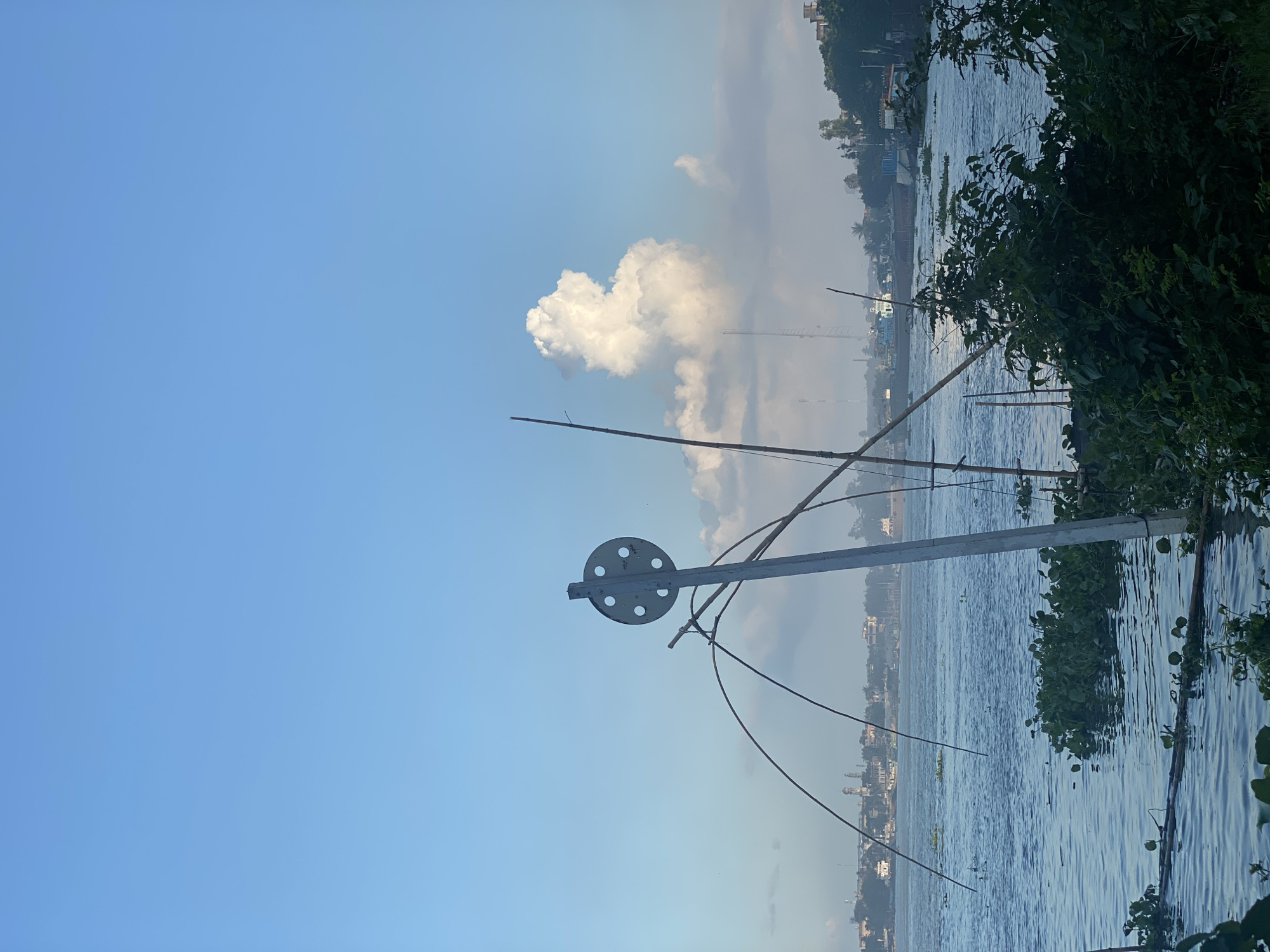 The image depicts a wide, tranquil river, likely the Buriganga River, as seen from the bank in Dhaka, Bangladesh. The sky is a clear, bright blue with a few scattered white clouds, one particularly large and fluffy cloud dominating the upper right portion of the frame. In the foreground, to the right, dense green foliage and reeds are visible, partially obscuring the view of the river.
Dominating the center of the image is a tall, slender pole with a circular, metallic attachment at the top. This attachment has several circular holes. Several long, thin bamboo poles extend outwards from the central pole, forming a structure that appears to be part of a fishing net or a similar apparatus. The water surface is calm, with subtle ripples. Patches of green water hyacinths float on the water.
In the middle ground, across the river, a cityscape is visible. Buildings, including what appear to be minarets or domes of mosques, are discernible along the shoreline. Several boats, including what looks like a larger cargo vessel, are present on the water. The distant landscape is somewhat hazy, suggesting distance and possibly a slight atmospheric diffusion. The overall impression is one of a serene riverside scene, typical of the outskirts of a bustling city like Dhaka, where nature and urban life coexist. There are no people visible in the frame.