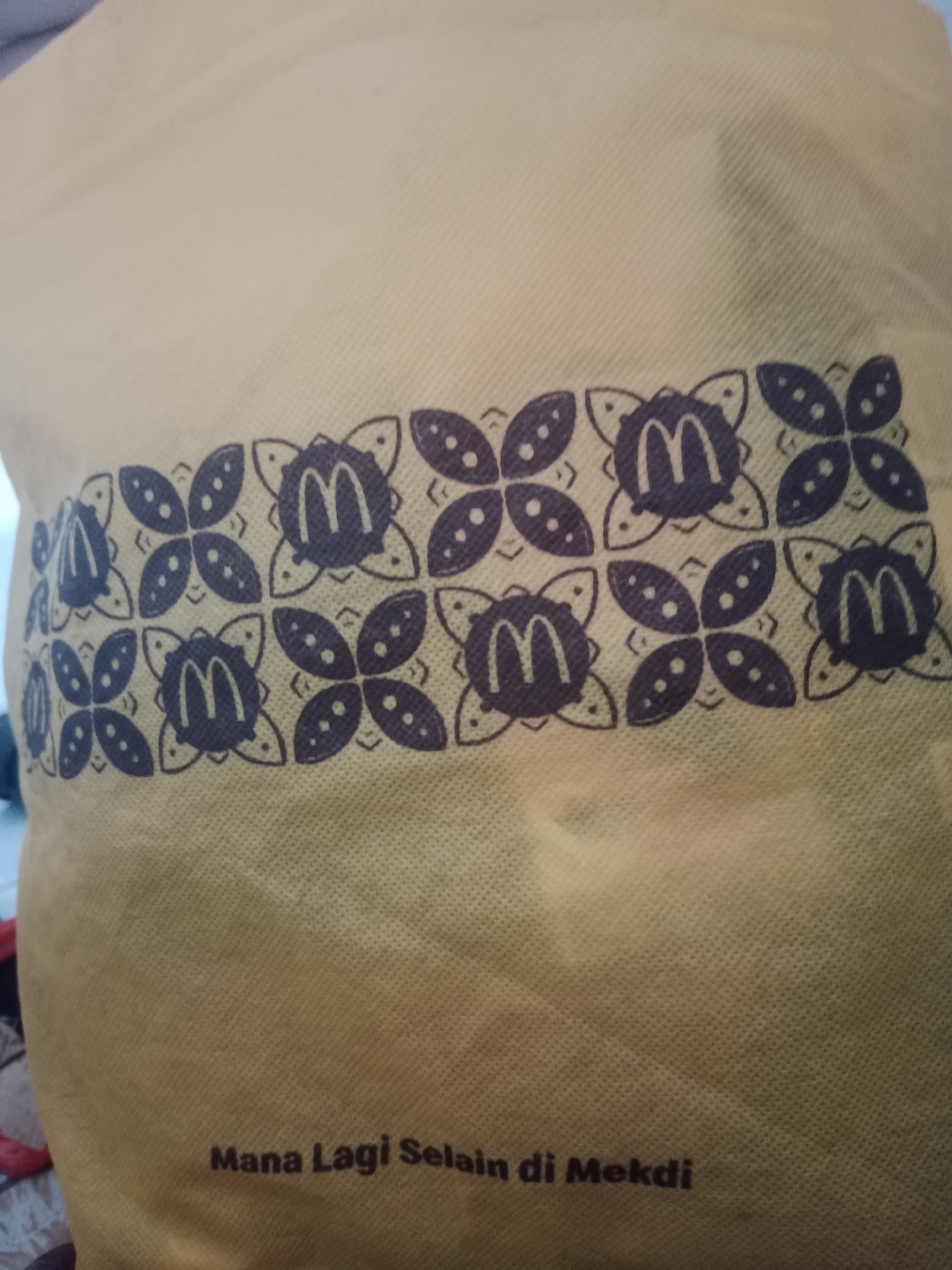 The image shows a close-up of a yellow fabric bag with a repeating pattern of stylized flowers and McDonald's "M" logos. The pattern is printed in dark blue. At the bottom of the bag, text in the same dark blue reads "Mana Lagi Selain di Mekdi". This phrase is Indonesian and translates to "Where else but at Mekdi?". "Mekdi" is a common colloquialism for McDonald's in Indonesia, particularly in Java. The background of the image is out of focus but appears to be an indoor setting with some light colored fabric visible, possibly part of the bag itself or other items. There are no people visible in the image. The image does not provide any information about the city of Yogyakarta, Indonesia.
