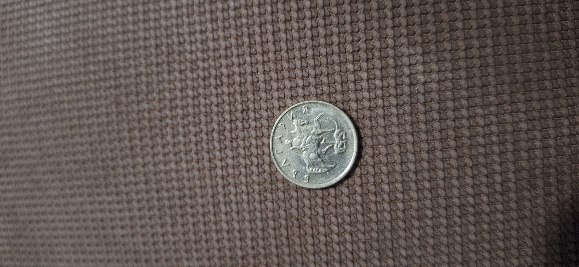 The image shows a Bulgarian 1 Lev coin from 1992 resting on a brown, textured surface. The coin is silver in color and depicts the Madara Rider. The surrounding area is out of focus. The location cannot be determined from this image.