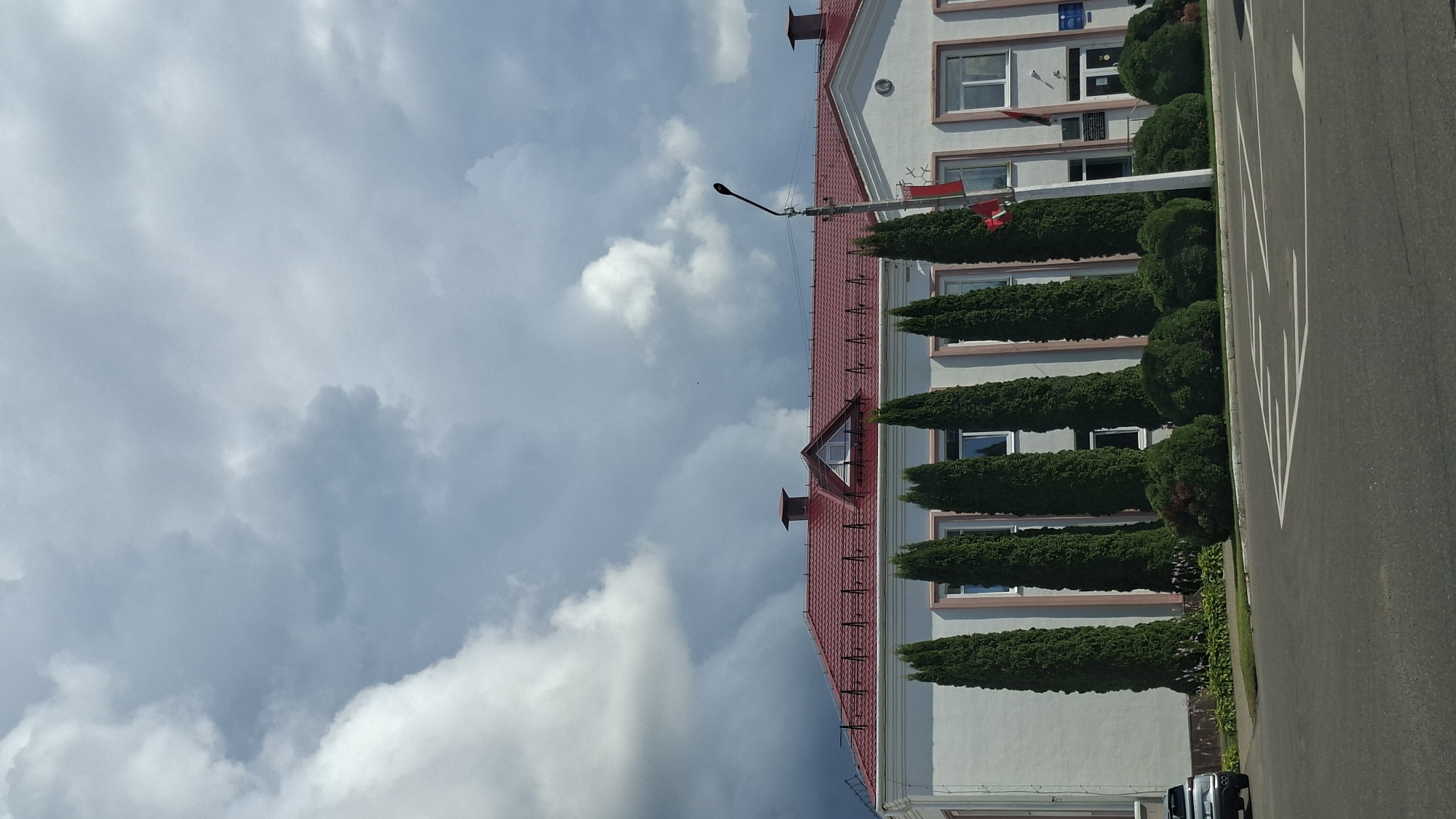 A wide-angle, vertical photograph captures the exterior of a modern administrative building with a prominent red roof, set against a dramatic sky filled with heavy, dark clouds. The building is light-colored, possibly white or cream, with several windows visible and decorative trim. A row of tall, slender evergreen trees, likely cypresses, stand in front of the building, interspersed with smaller, rounded bushes. A street with white directional arrows painted on the asphalt occupies the foreground. A dark-colored car is parked on the left side of the road, facing away from the camera. A lamppost with a single light fixture is positioned to the right of the trees, near the building's entrance. Two flags are visible on flagpoles attached to the building. The overall impression is one of an institutional setting under an impending storm. This scene is likely located in the city of Myadzyel, Belarus, given the architectural style and the presence of national or regional flags.