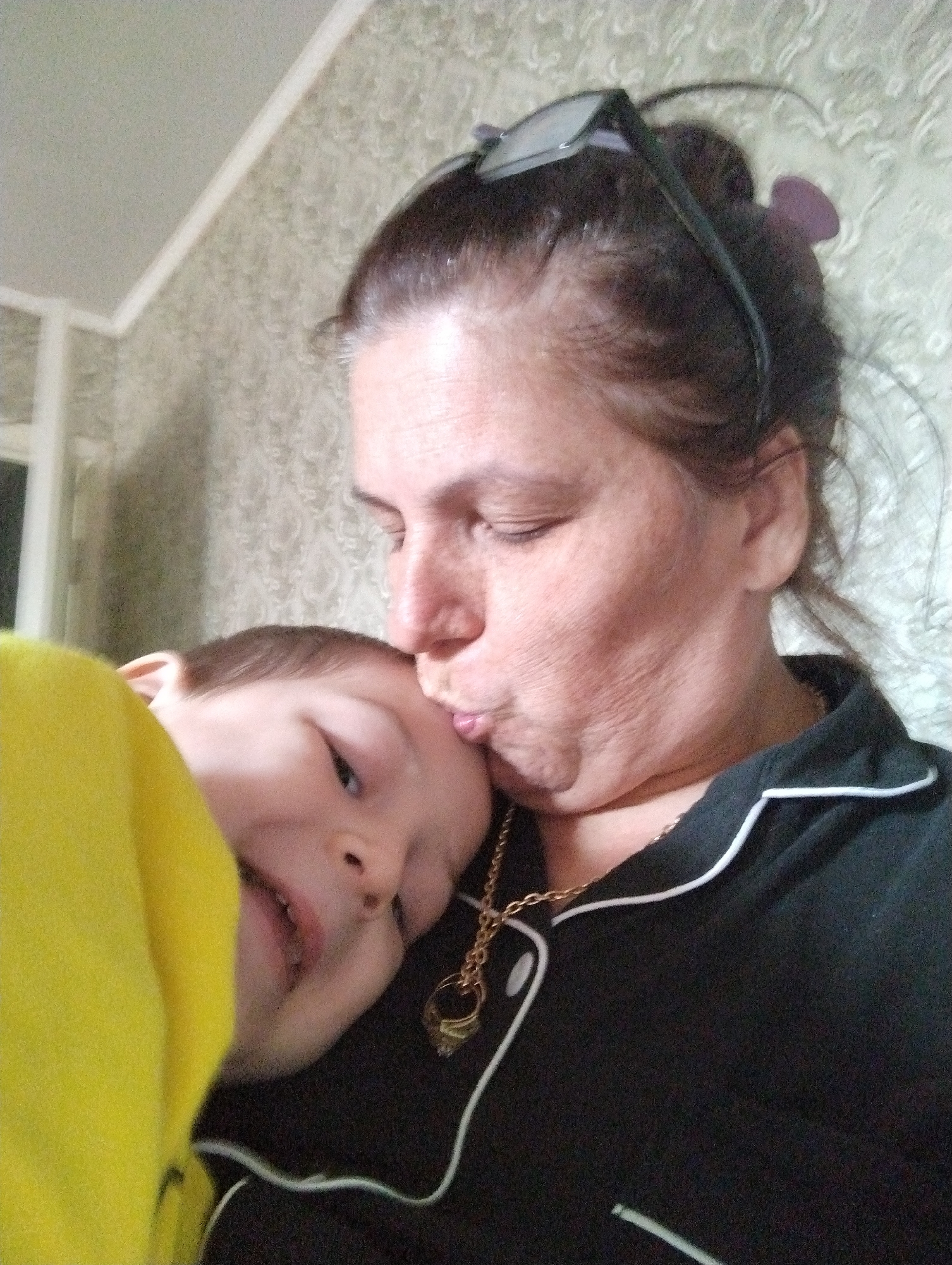 A woman is kissing a young child on the forehead. Both are in focus. The woman has dark hair pulled back and is wearing black pajamas with white trim and a gold chain necklace. The child, wrapped in a bright yellow blanket, is looking at the camera. The background is a wall with textured wallpaper. The lighting is dim and appears to be from an indoor source. The overall impression is one of an intimate moment between a caregiver and child. The scene is likely indoors, possibly in a home. There are no specific landmarks or clues to pinpoint the location to Kingisepp, Russia.