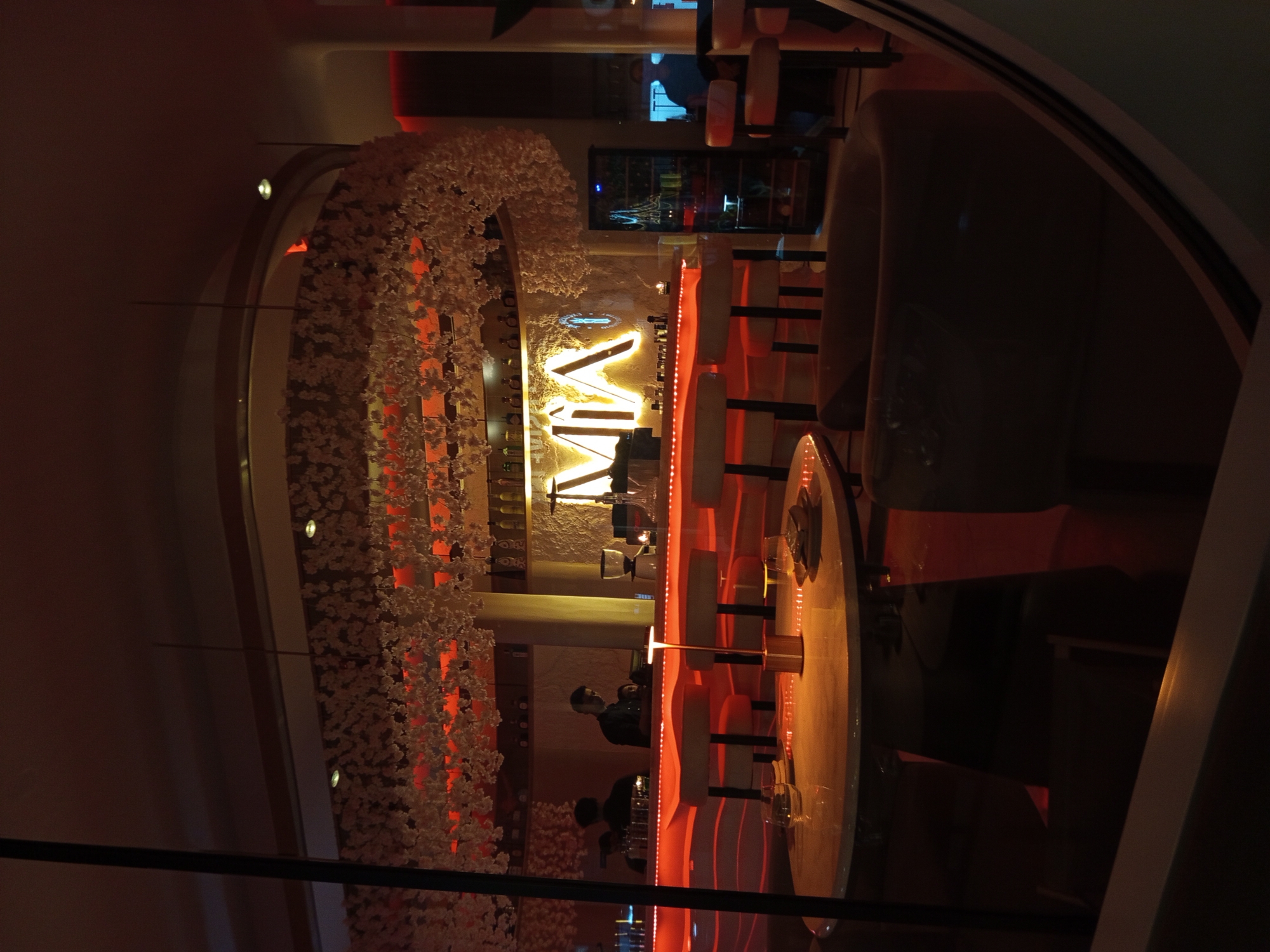 The image depicts the interior of a bar, likely in North Kuta, Indonesia, characterized by a modern and decorative style. The bar area features high stools, a round table, and a lit-up logo on the wall that says "VIA". A large, floral arrangement hangs above the bar. There are a few people present, including a bartender behind the bar. The lighting in the space is dim, with red-orange accents highlighting the bar area. A glass or reflective surface appears in the foreground, partially obscuring the view.