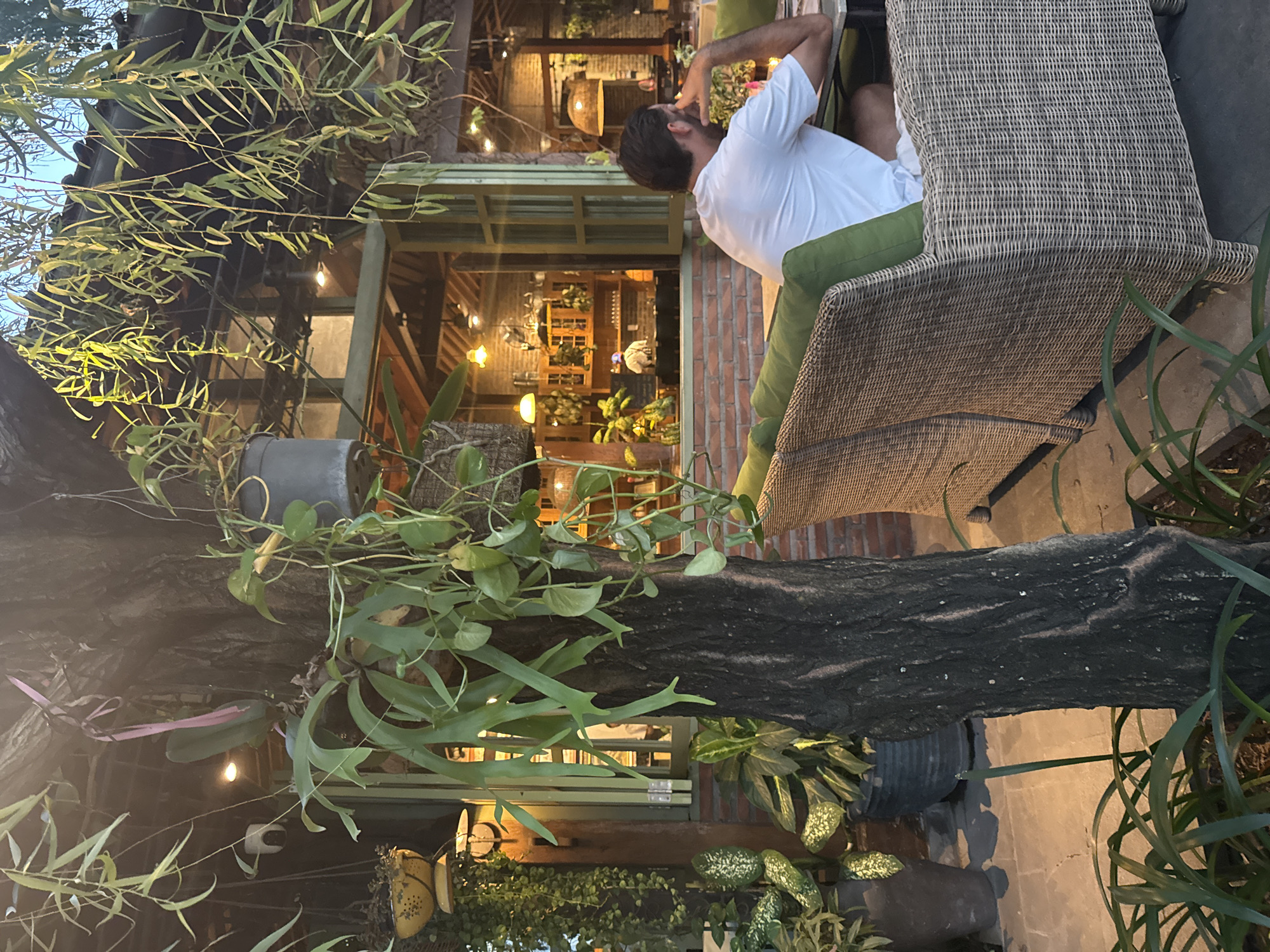 The image depicts an outdoor seating area, likely in North Kuta, Indonesia, given the tropical vegetation and architectural style. A man wearing a white shirt is seated in a wicker chair with green cushions. The chair is positioned outdoors. The background reveals an interior space with warm lighting, brick walls, and shelves displaying various items. Lush greenery, including hanging plants and trees, surrounds the seating area, creating a natural atmosphere. A thick tree trunk with climbing plants stands in the foreground. The lighting suggests it is evening.