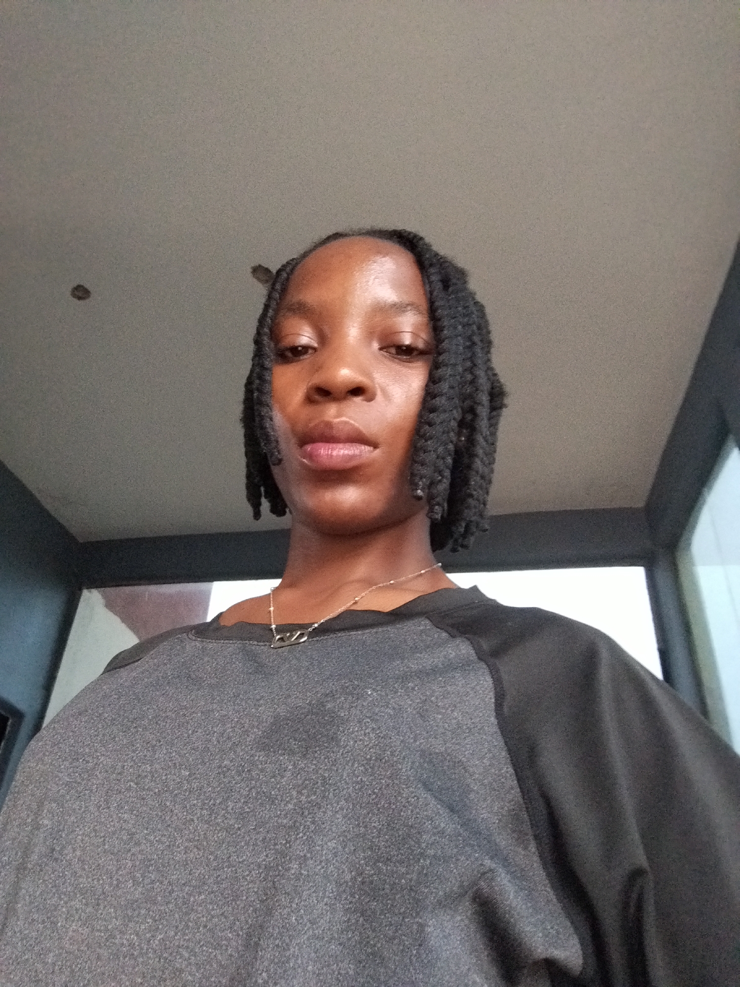 The image shows a medium shot of a young Black woman from the chest up, looking directly at the camera. She has her hair braided into dreadlocks that frame her face. She is wearing a grey shirt with black sleeves and a silver necklace with a pendant. The background appears to be indoors, with a grey ceiling and a darker wall or structure behind her. The overall lighting is somewhat dim. The location is Uselu, Nigeria.