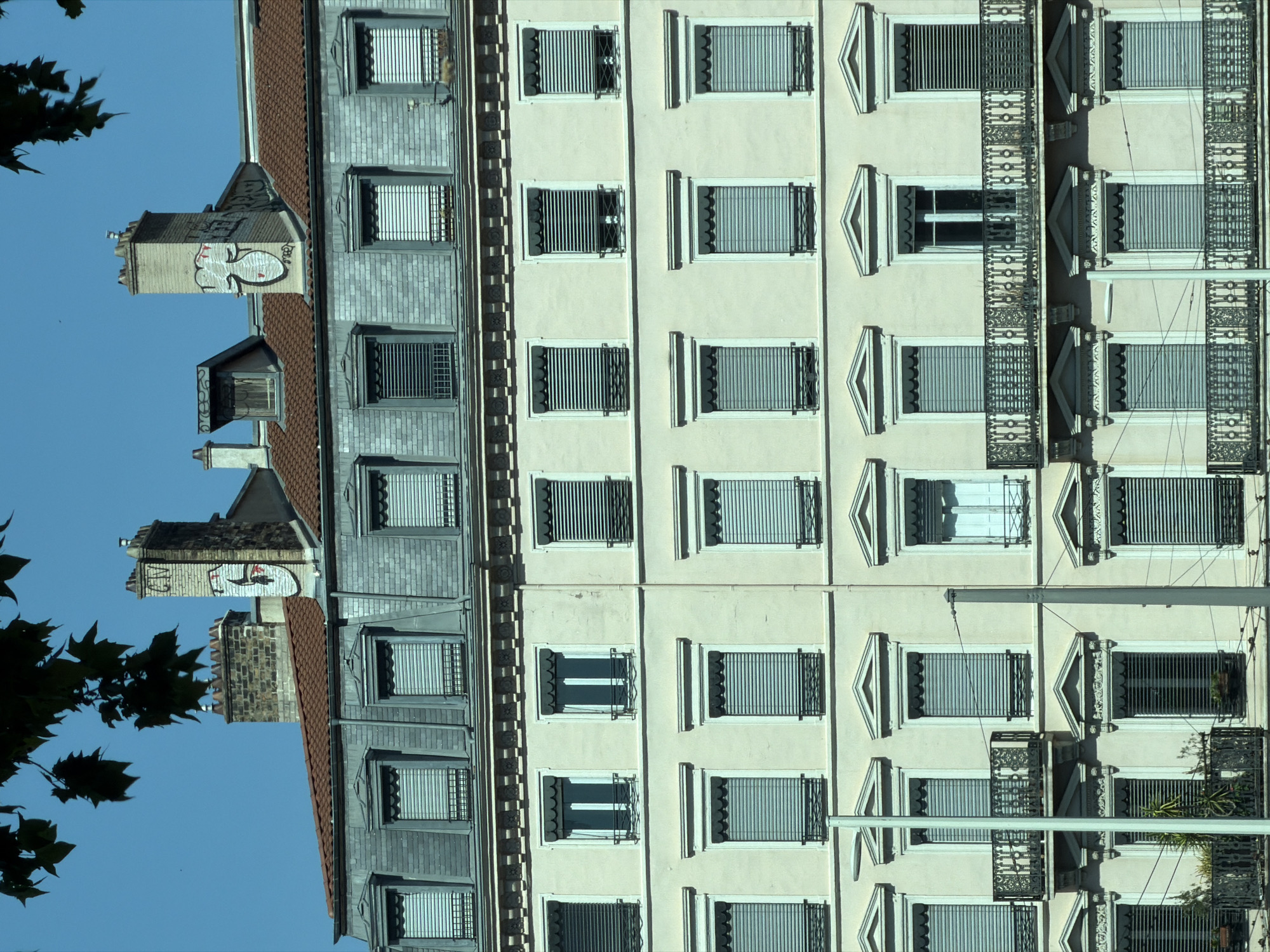 The image shows the facade of a building in Lyon, France, with multiple floors of windows and balconies. The building's exterior is a pale yellow color. The windows are mostly covered by horizontal blinds, and some have decorative awnings. There are two balconies visible, featuring ornate metal railings. The roofline is sloped and covered in grey tiles, with dormer windows and chimneys. Two of the chimneys are decorated with graffiti. The sky is clear and blue. Dark green leaves from a tree hang from the top left corner of the image. The image appears to be taken from a street level perspective, looking upwards at the building.
