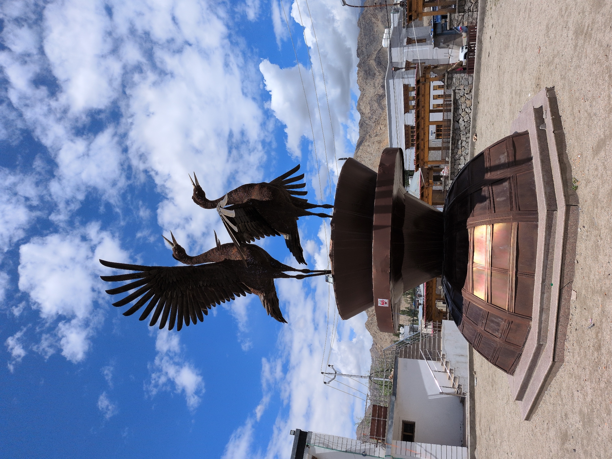 The image shows a bronze sculpture of two cranes with their wings spread, standing on a tiered, dome-shaped pedestal. The sculpture is located in an outdoor area with a sandy ground and a backdrop of buildings and mountains. The sky is blue with scattered white clouds. In the foreground, to the left, is a white building with a staircase leading up to a balcony. To the right, there are more buildings with wooden architectural elements. The mountains in the background are dry and rocky. The overall impression is of a public art installation in a high-altitude, possibly arid region. The specific location is in the city of Leh, India, as indicated by the architectural style of the buildings and the surrounding landscape.