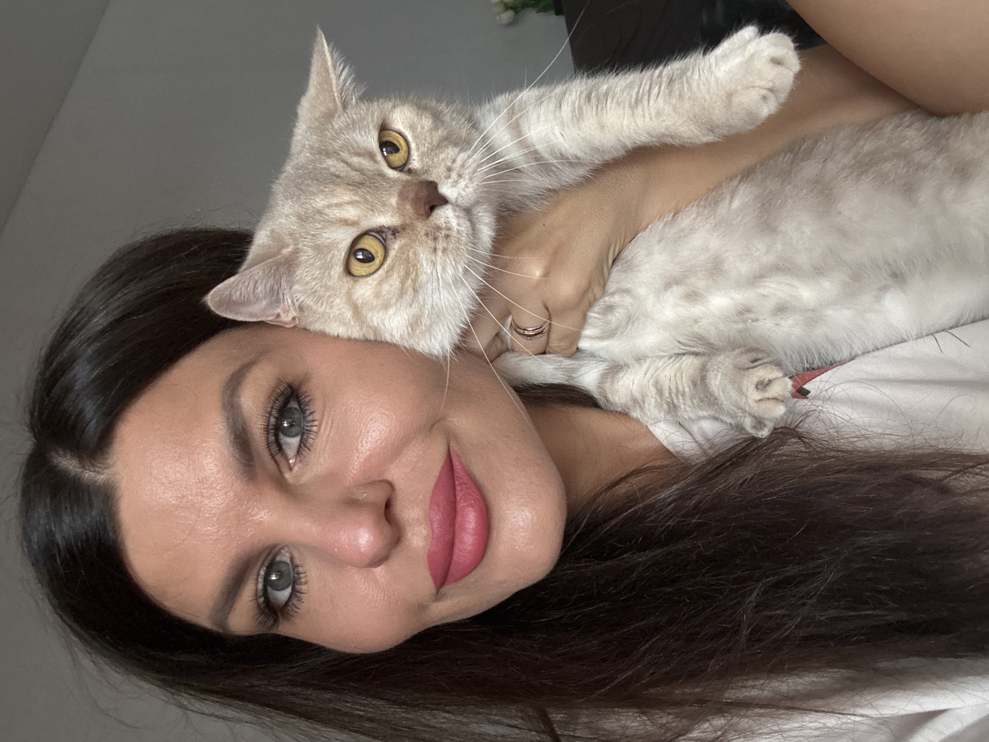 A woman with dark hair and full lips, wearing pink lipstick, is holding a light-colored cat. The cat has yellow eyes and is looking at the camera. The woman's hair is dark and appears to be styled naturally. The background is a neutral gray color, suggesting an indoor setting. There is no identifiable information in the image to suggest a specific location in Chaikovsky, Russia.