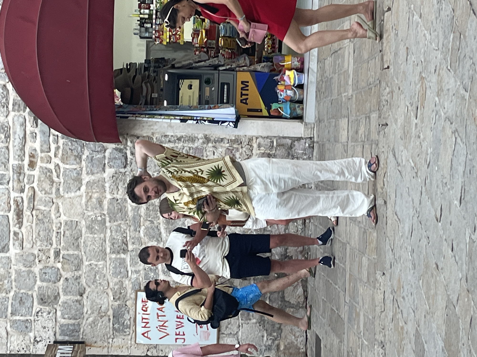 A group of people are standing on a cobblestone street in front of a building with a stone facade. A red awning is visible above a shop entrance. In the foreground, a man in a patterned shirt and white pants is holding what appears to be a pineapple and a phone. To his left, a man in a white t-shirt and dark shorts is looking at his phone. Further to the left, another man in a tan shirt and blue shorts is also looking at his phone. A woman in a red dress is walking away from the viewer to the right. An ATM machine is visible inside the shop. The street appears to be in the Old Town of Budva, Montenegro, given the architecture and the presence of tourist-oriented shops.