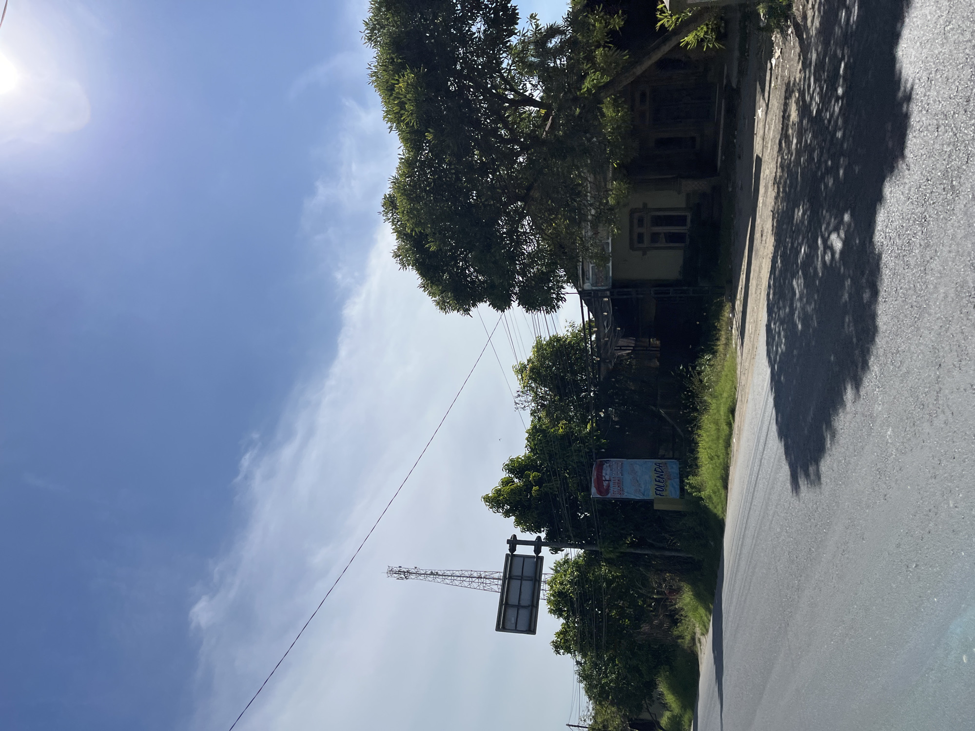 The image depicts a roadside scene in Bukitrata, Indonesia, under a clear blue sky with scattered clouds. The sun is bright, suggesting it is daytime, likely midday.
In the foreground, a paved road stretches into the distance, with the shadow of a tree cast across its surface. To the left of the road, there's a narrow strip of green grass and vegetation. Beyond the grass, a building with light-colored walls and a dark roof is visible, partially obscured by the lush foliage of a large tree. The building appears to have windows with decorative frames.
Further down the road, a cellular tower stands tall, indicating a developed area with communication infrastructure. Next to the tower, a rectangular sign is mounted on a pole, displaying text that appears to be in Indonesian. One visible word on the sign is "FOLENDA". Another smaller sign, possibly an advertisement, is also present, featuring a picture and some text.
The overall scene conveys a sense of a rural or semi-urban environment in Indonesia, characterized by natural greenery, local architecture, and modern telecommunications. The bright sunlight and clear sky suggest pleasant weather conditions. No people are visible in the image.