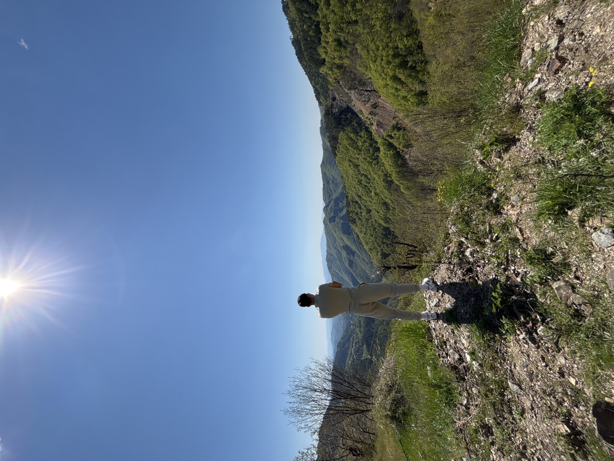 The image shows a man standing on a rocky, grassy ridge overlooking a mountainous valley in Petrov Dol, Bulgaria. The man is facing away from the camera, wearing light-colored clothing. The sky is clear blue with the sun visible in the upper left corner. The valley is densely forested with green trees. The ridge is composed of rocks, dirt, and grass.