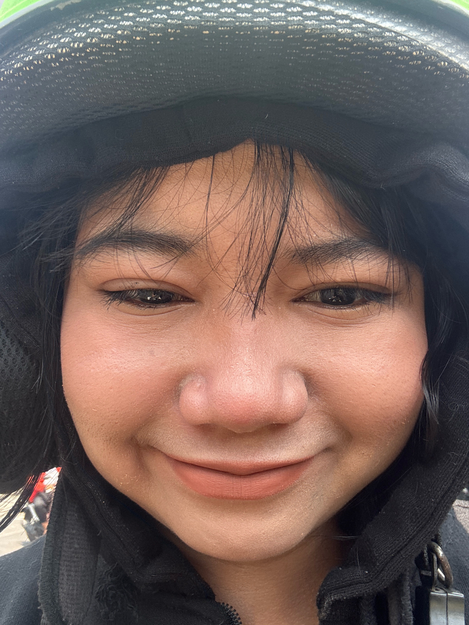 A close-up, front-facing selfie shows a person wearing a black helmet with a light green brim and a black hooded jacket. The person's face is mostly visible, with dark hair framing their face. They are wearing light brown lipstick and subtle eye makeup, including eyeliner. The background is blurred, but it appears to be an outdoor setting, possibly in North Jakarta, Indonesia, with hints of other vehicles visible. The lighting suggests it is daytime.