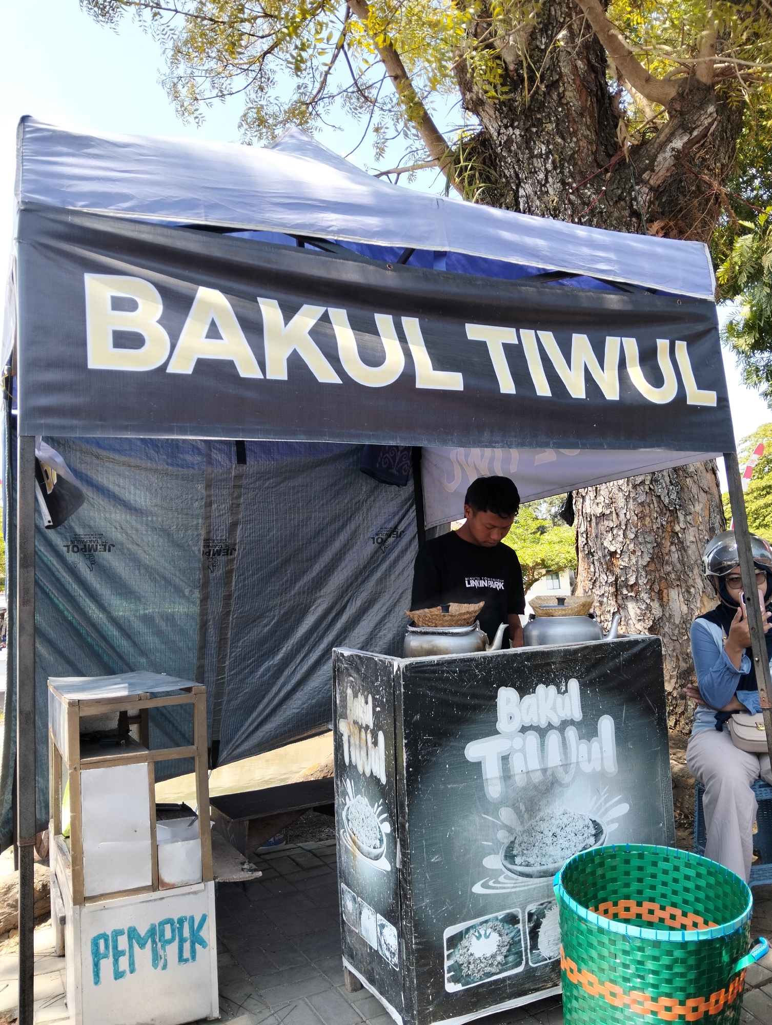 In Magelang, Indonesia, a street vendor stall operates under a large tent. The stall displays a prominent banner reading "BAKUL TIWUL". A young man in a black t-shirt is seen behind a counter, appearing to be preparing food. On the counter, there are two large metal pots with woven baskets placed on top. To the left of the stall, a smaller cart is labeled "PEMPEK". To the right of the stall, a woman wearing a helmet is seated, holding a mobile phone. The stall is situated outdoors, with a large tree providing shade and a clear blue sky overhead. The ground is paved with interlocking bricks.