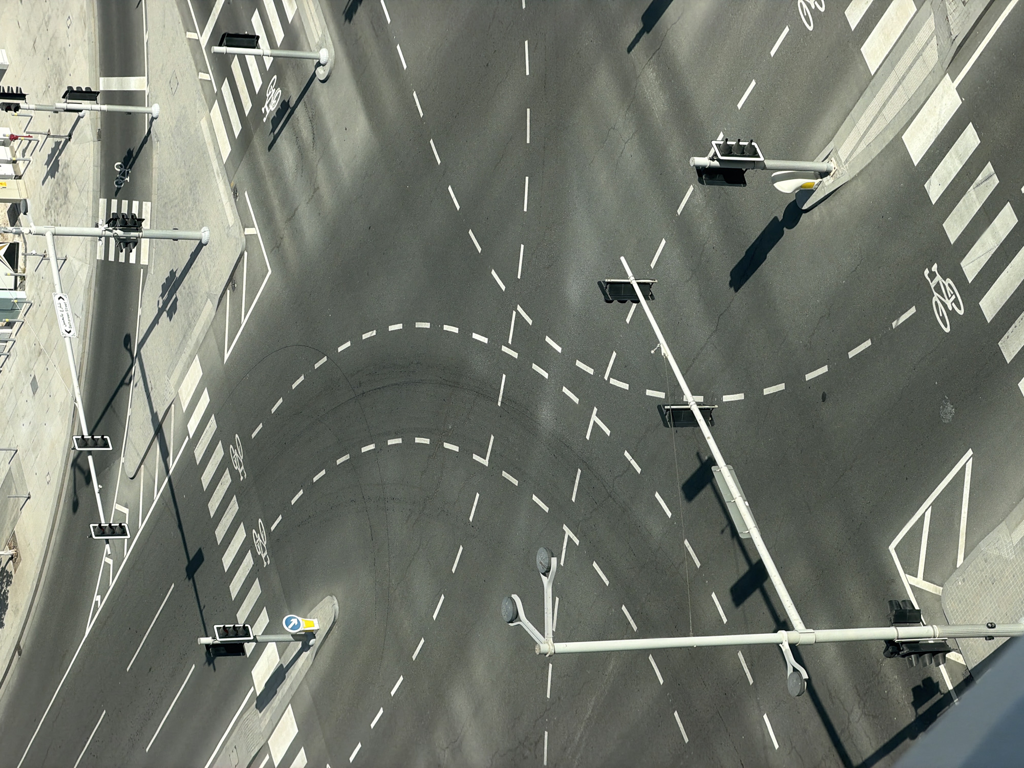 This is an aerial, high-angle, eye-level shot of an empty intersection in a city, likely during daylight hours due to the distinct shadows cast by the traffic lights and street lamps. The intersection features multiple lanes of asphalt road with clear white lane markings, including solid and dashed lines, as well as pedestrian crosswalks. Several traffic light poles with black signal heads are visible, some accompanied by street lamps. There are also directional signs, including a blue and yellow sign with a white arrow pointing left and a white street sign with Arabic and English text, which reads "Corniche Street." Bicycle lane markings are present on some of the crosswalks, depicting a bicycle symbol. The intersection appears to be at an urban crossroads, with paved sidewalks and some grey, textured pavement around the edges. No vehicles or people are present in the scene. The overall impression is one of an orderly, well-maintained urban infrastructure.