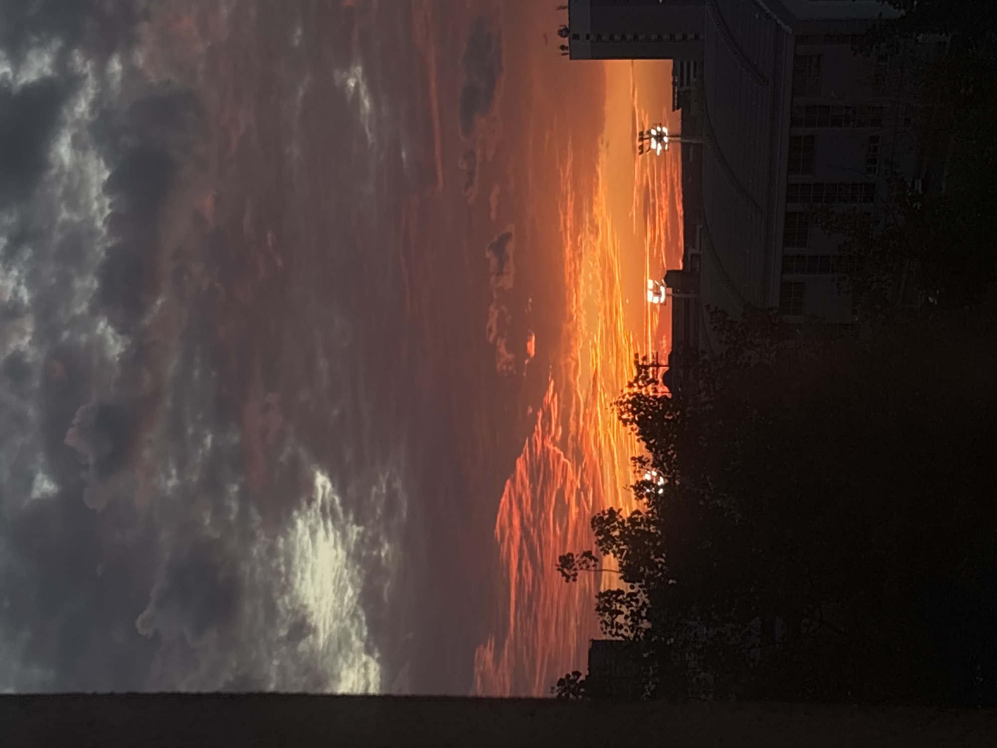 The image shows a sunset scene in Minsk, Belarus. To the left, heavy, dark clouds with some illuminated sections fill a significant portion of the sky. Moving rightward, the clouds transition into a vibrant orange and red hue, typical of a sunset. The sun is obscured by buildings and trees. The silhouette of a building structure is visible to the right. Below the horizon, the foreground is dark with the outline of trees. The bottom of the image shows a retaining wall of some kind.