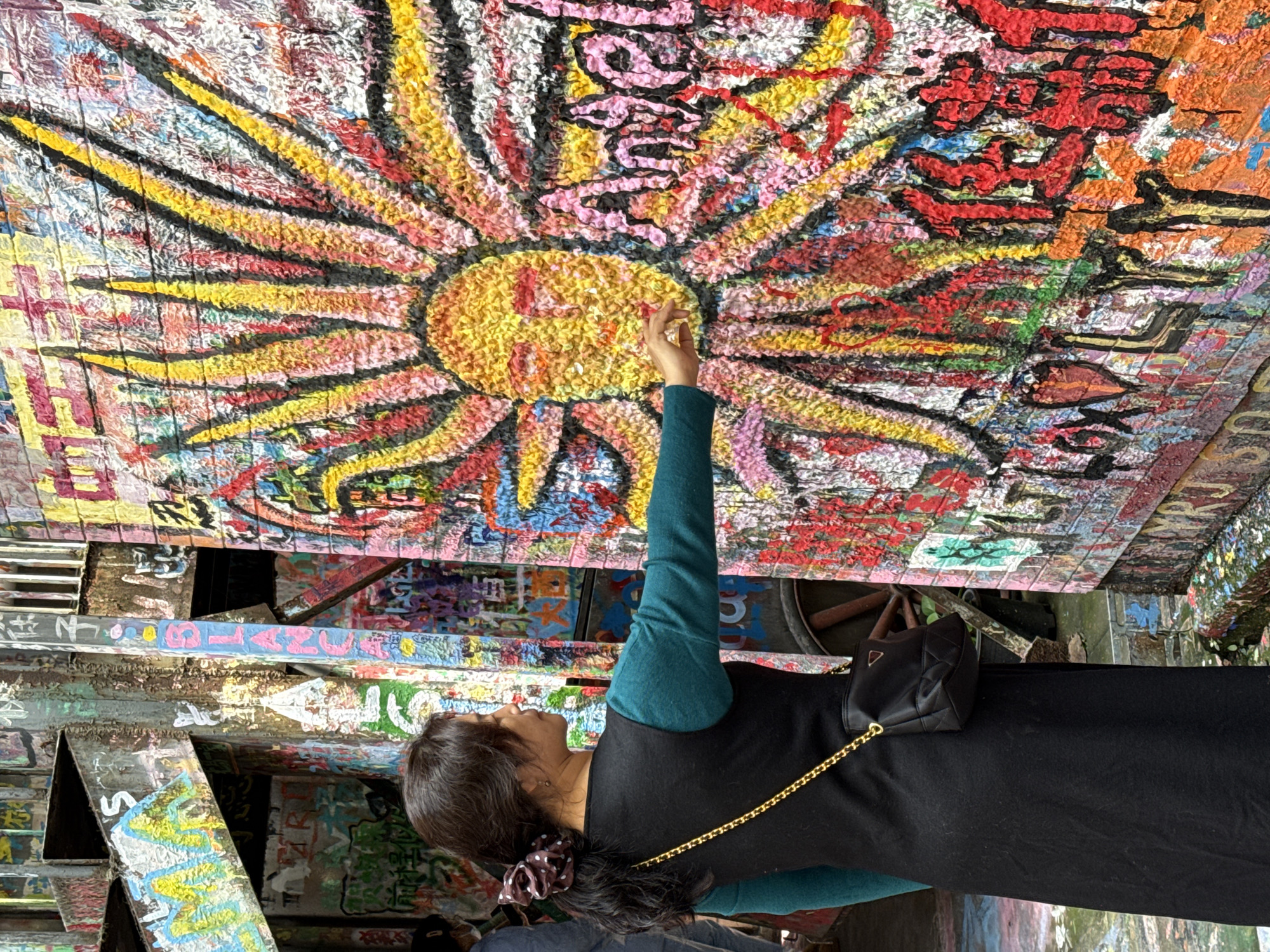 The scene is set in a graffiti-covered alleyway in Jiulongpo District, China. A female individual, seen from a three-quarter rear perspective, occupies the mid-ground. She has dark hair styled in a ponytail secured with a dark brown polka-dotted scrunchie. She wears a long-sleeved dark teal top under a black sleeveless long dress and carries a black quilted shoulder bag with a gold chain strap. Her right arm is extended, with her index finger making contact with a large, textured sun graffiti artwork on the adjacent wall. This prominent artwork features a yellow facial area with pink details and radiating rays in yellow, red, black, and pink, all rendered in a thick, almost sculptural paint application. To the right of the sun, the word "Angel" is painted in pink. All visible wall surfaces and structural components are heavily covered with vibrant, multi-layered graffiti, including various tags, Chinese characters, and abstract designs. In the far background to the left, a partially visible individual wearing a face mask is discernible. The ground appears to be damp or reflective.