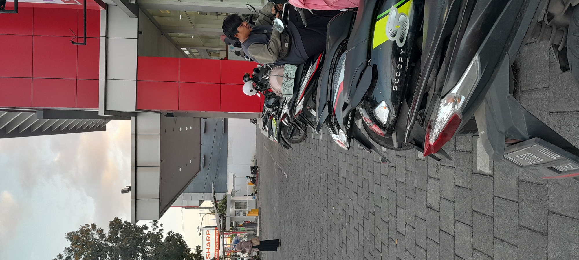 An exterior shot captures a street scene in Rogojampi, Indonesia, with a prominent modern building featuring red and silver panels. In the foreground, several motorcycles are parked on a paved lot. One man is seated on a motorcycle, looking at his phone. To the left, a woman wearing a hijab stands holding a bag. A sign for "SHARP ELECTRONICS" is visible in the background. The sky is overcast with some clouds.