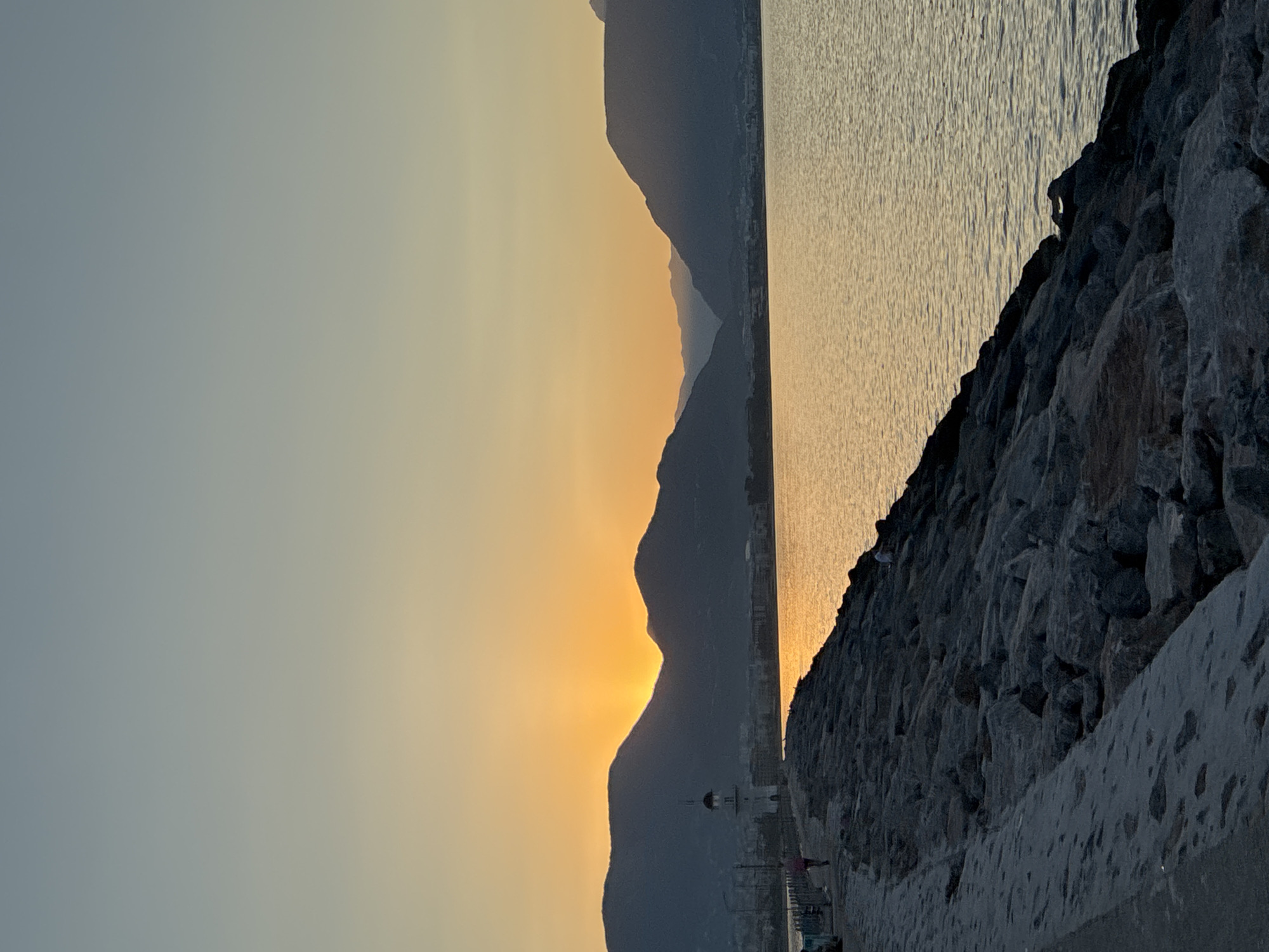 The image depicts a sunset over the sea in Alanya, Turkey. In the foreground, a rocky breakwater extends into the water, with a paved path running alongside its left edge. A lighthouse is visible on the breakwater, and a person is walking along the path. In the middle ground, the sea reflects the warm colors of the sunset, and a city skyline can be seen along the coast. The background features silhouetted mountains under a gradient sky transitioning from yellow near the horizon to pale blue at the top.