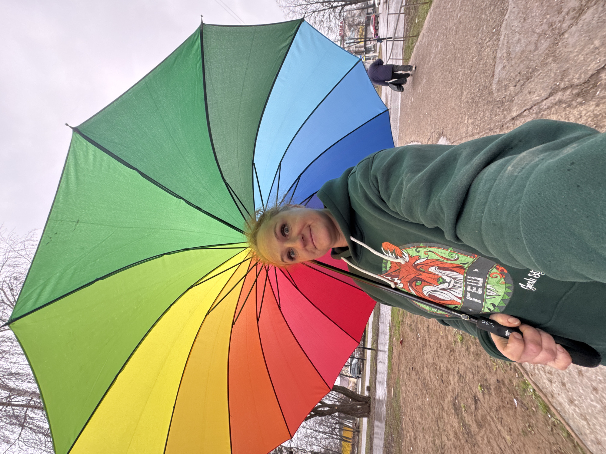 A woman is holding a rainbow umbrella while taking a selfie outdoors. She is wearing a green hooded sweatshirt with a graphic design. In the background, there is a sidewalk, street, and buildings. Bare trees are visible. This image was taken in Chaikovsky, Russia.