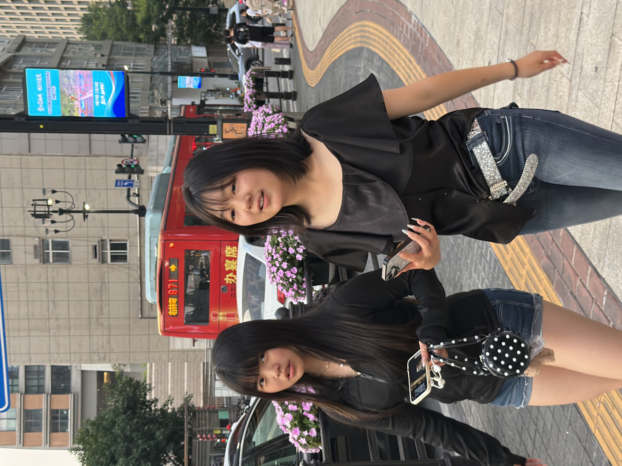 This image captures two East Asian women walking on a paved urban sidewalk in Yuzhong District, China.
The woman on the left has long, straight, black hair with bangs, and is looking slightly to her left with a slightly parted mouth. She is wearing a black long-sleeved zip-up top and blue denim cut-off shorts. She wears multiple layered gold necklaces. In her left hand, she holds a silver smartphone with white wired earbuds dangling from it, and a small, circular black bag with white polka dots and a brown plush charm. Her fingernails are long and pointed.
The woman on the right has short, straight, black hair, chin-length, and is looking forward and slightly to her right with a faint smile. She is wearing a black short-sleeved top with a ruffled overlay on the bodice, dark blue denim jeans, and a prominent wide silver glittery belt with a large buckle. In her left hand, she holds a silver smartphone. A dark bracelet is visible on her left wrist. Her fingernails are long, pointed, and light-colored.
Both women appear to be walking in parallel, suggesting they are together. The sidewalk features a yellow tactile paving strip.
In the background, a red public bus with the number "871" and Chinese characters "渝中" (Yuzhong) and "办宴席" (host a banquet) is visible. Behind the bus, a white car and a gray car are parked or moving. Multi-story buildings with windows are present on both sides of the street. Streetlights, traffic lights (displaying red and green signals), and digital billboards with blue and white displays showing Chinese text and images are also visible. Pink flowering plants are arranged in planters along the sidewalk. Additional blurry figures, including one pushing a stroller, are present in the distant background. The lighting suggests daytime, possibly late afternoon.